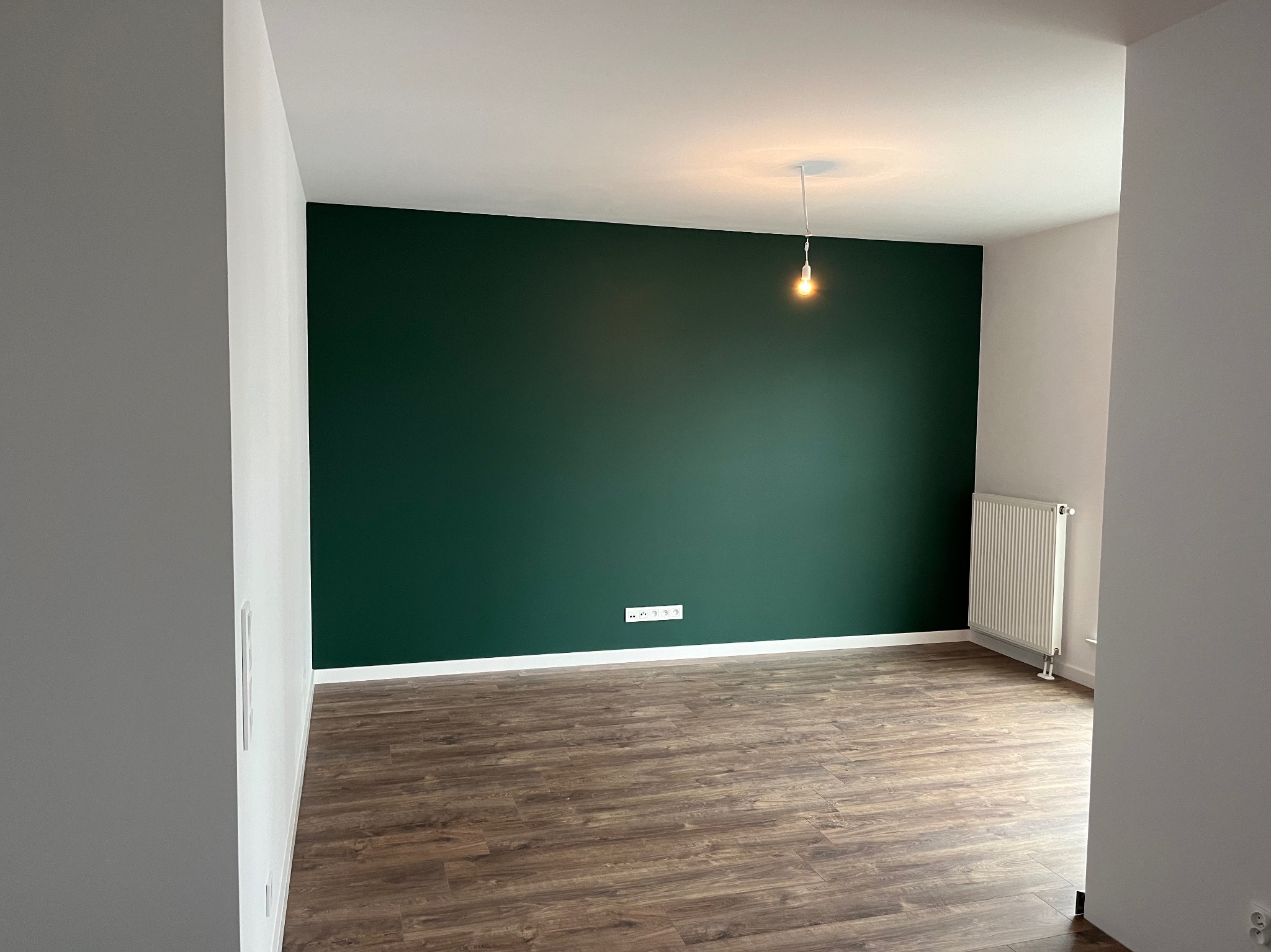 Pomalowany na ciemnozielono pokój z drewnianą podłogą, białymi listwami przypodłogowymi i grzejnikiem, oświetlony pojedynczą żarówką na kablu.