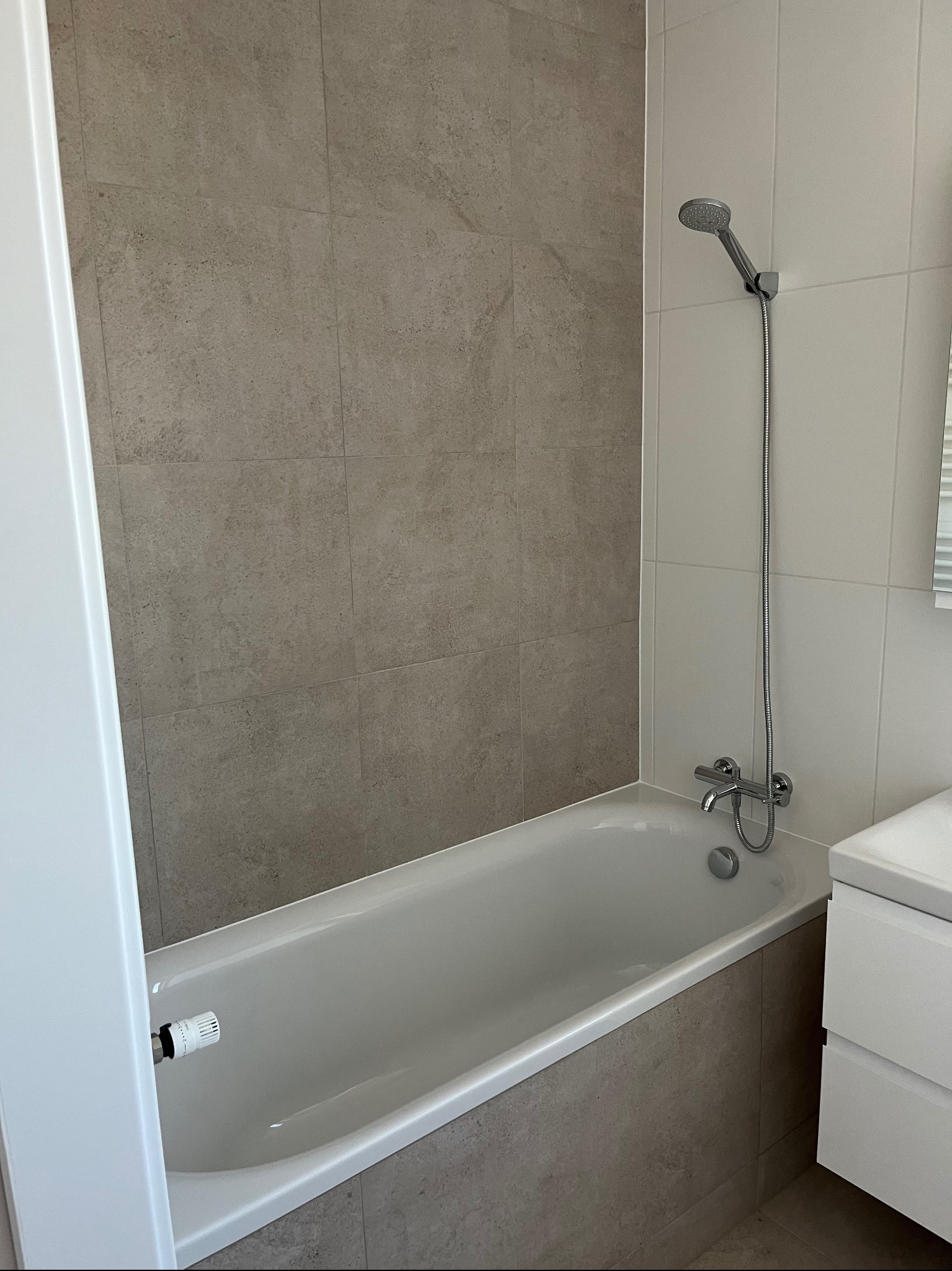 Wykończona łazienka w Poznaniu: beżowe płytki imitujące kamień na ścianach i obudowie wanny, biała wanna z baterią i słuchawką prysznicową, biała szafka pod umywalką.