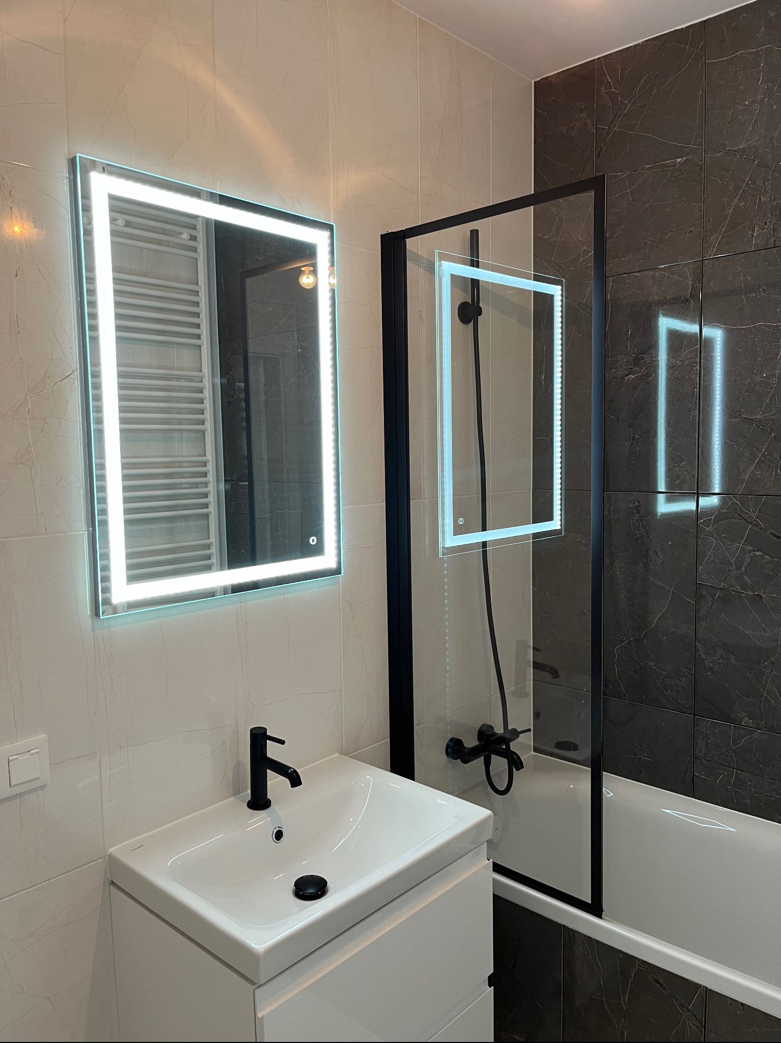 Nowoczesna łazienka z podświetlanym lustrem, czarną armaturą i kabiną prysznicową typu walk-in z czarnymi profilami na tle płytek imitujących marmur.
