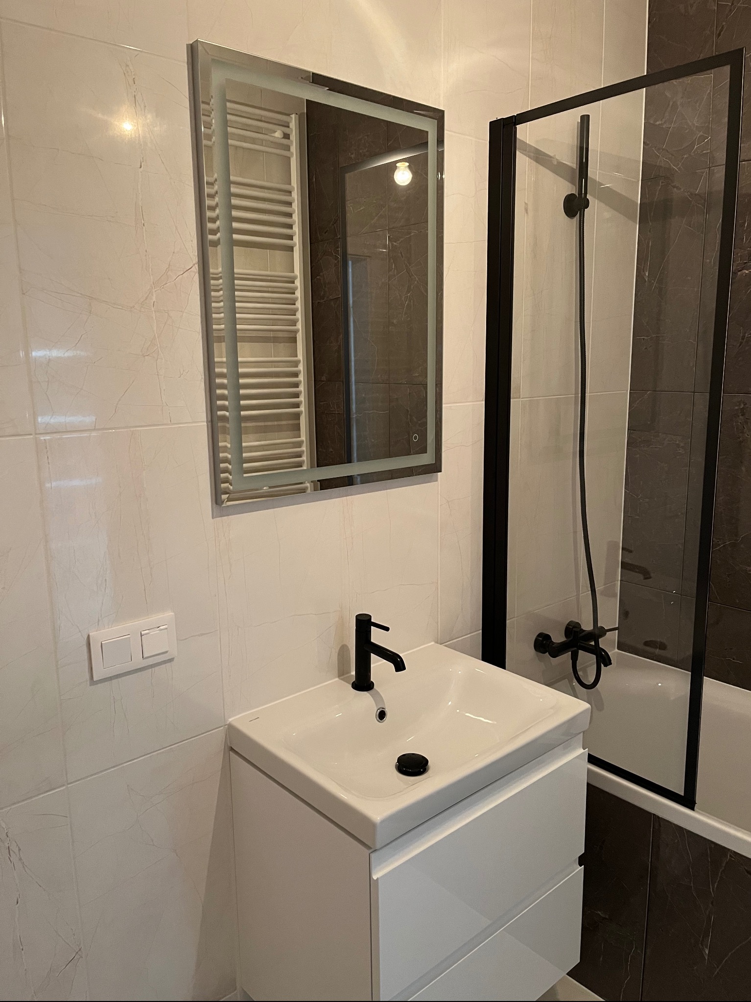 Nowoczesna łazienka z umywalką na szafce, czarną armaturą i kabiną prysznicową z czarnymi profilami, widok na marmurowe płytki ścienne i podświetlane lustro.