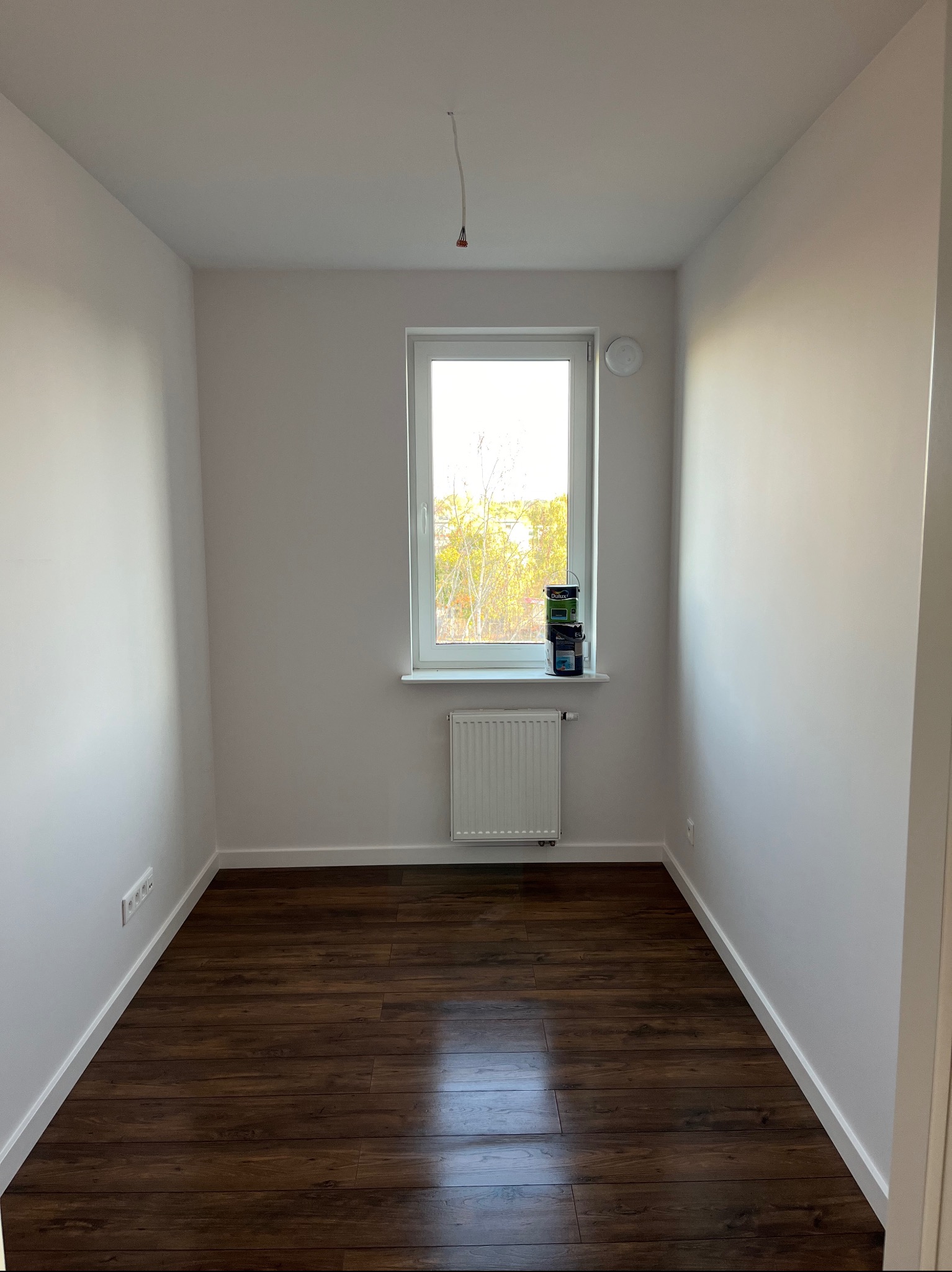 Pusty pokój po remoncie z brązową podłogą z paneli, białymi ścianami i grzejnikiem pod oknem, na parapecie stoją puszki z farbą.