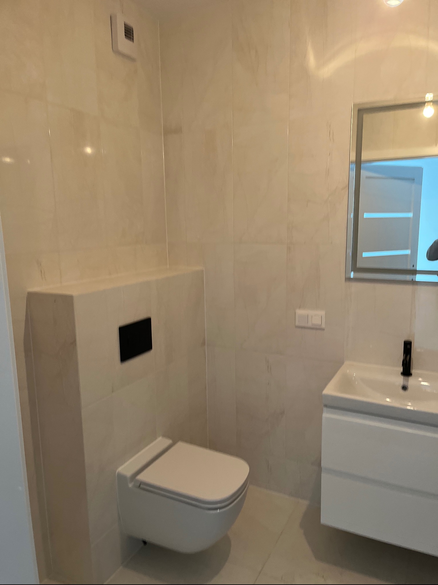 Nowoczesna łazienka z białymi płytkami imitującymi marmur, podwieszana toaleta z czarnym przyciskiem, biała szafka z umywalką i czarną baterią oraz lustro z oświetleniem LED