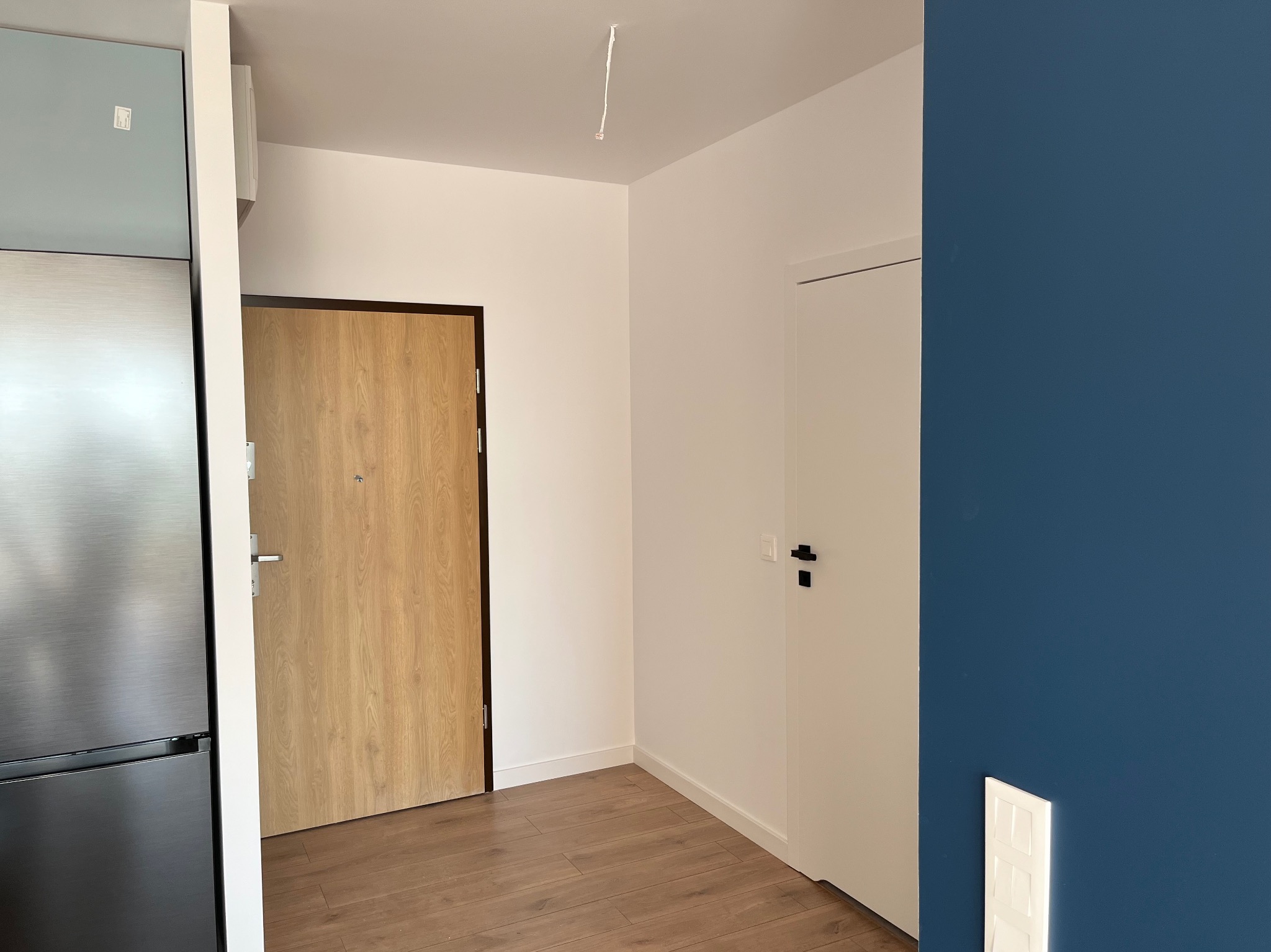 Nowoczesne wnętrze mieszkania po remoncie: drewnopodobne panele podłogowe, białe ściany, dwoje drzwi (jedne drewniane, drugie białe), fragment lodówki i ściana pomalowana na granatowo.