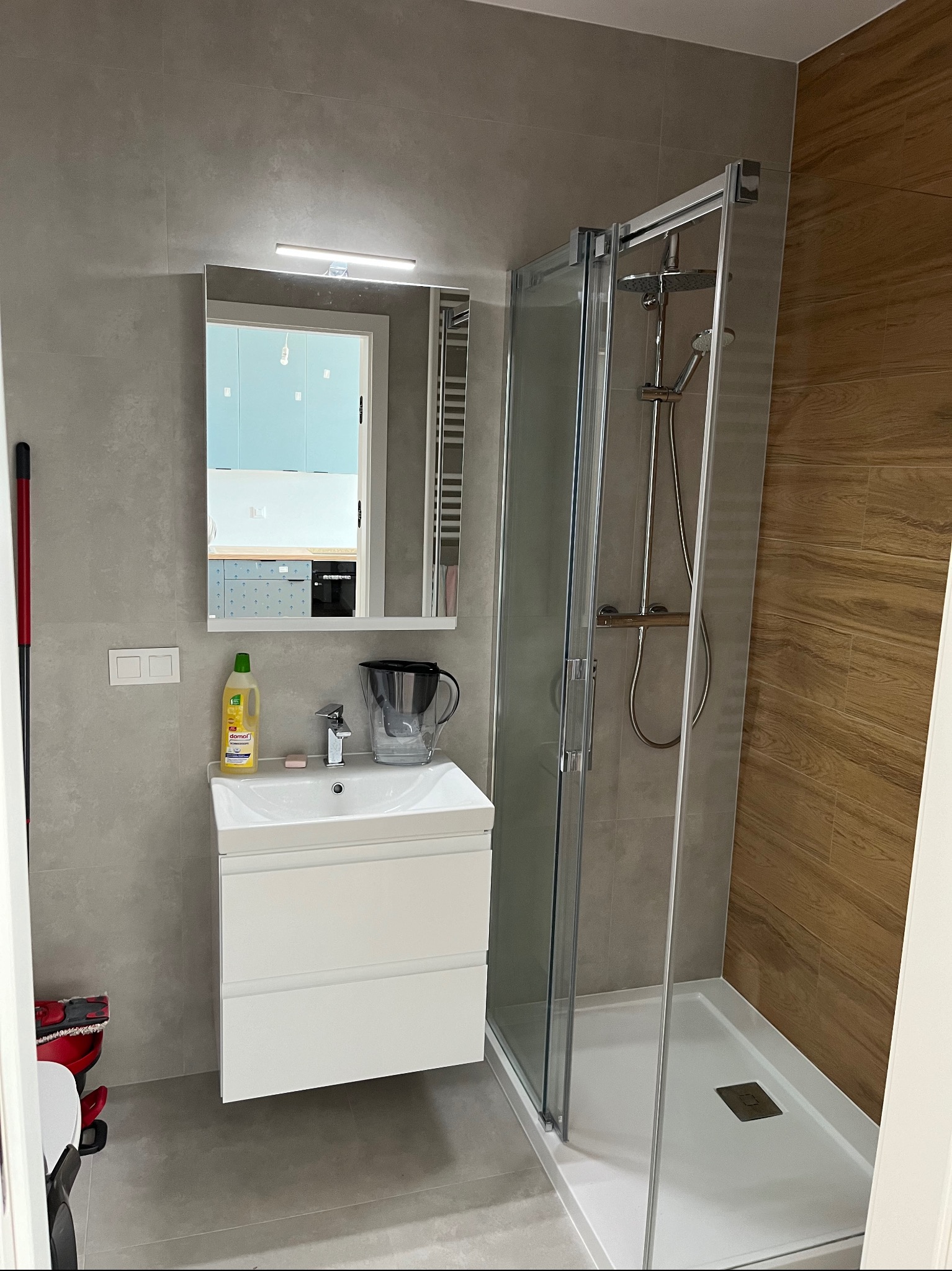 Nowoczesna łazienka po remoncie: kabina prysznicowa z chromowaną armaturą, biała umywalka z szafką, lustro z oświetleniem nad umywalką, szare płytki na ścianach i podłodze, oraz drewnopodobne...