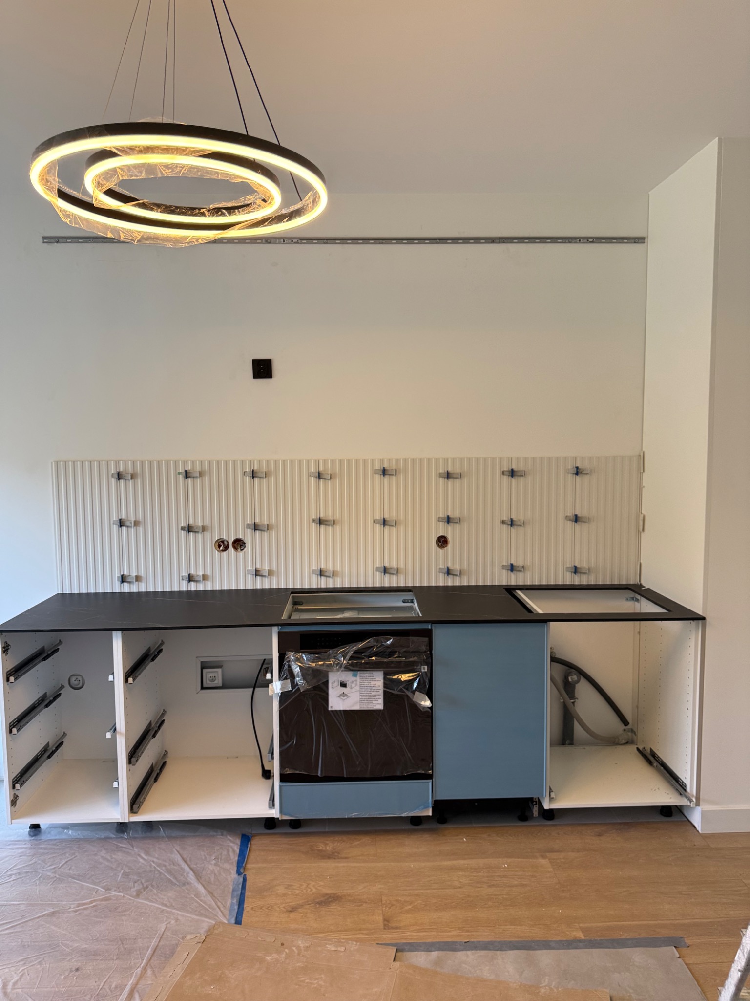 Montaż mebli kuchennych: widok na szafki bez frontów, blat z wycięciem na zlew i fragment ułożonej, białej mozaiki ściennej z krzyżykami dystansowymi.