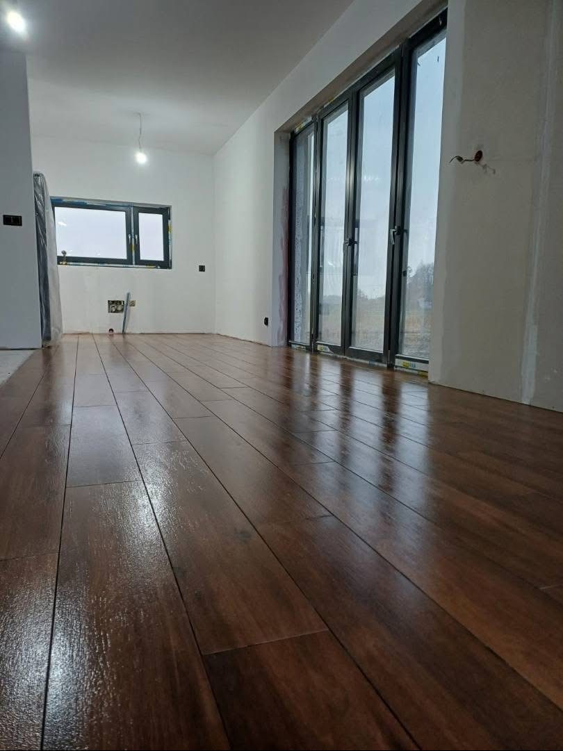 Błyszcząca, świeżo ułożona podłoga z paneli imitujących ciemne drewno w jasnym wnętrzu z dużymi oknami balkonowymi i standardowym oknem. Widoczne niedokończone ściany.