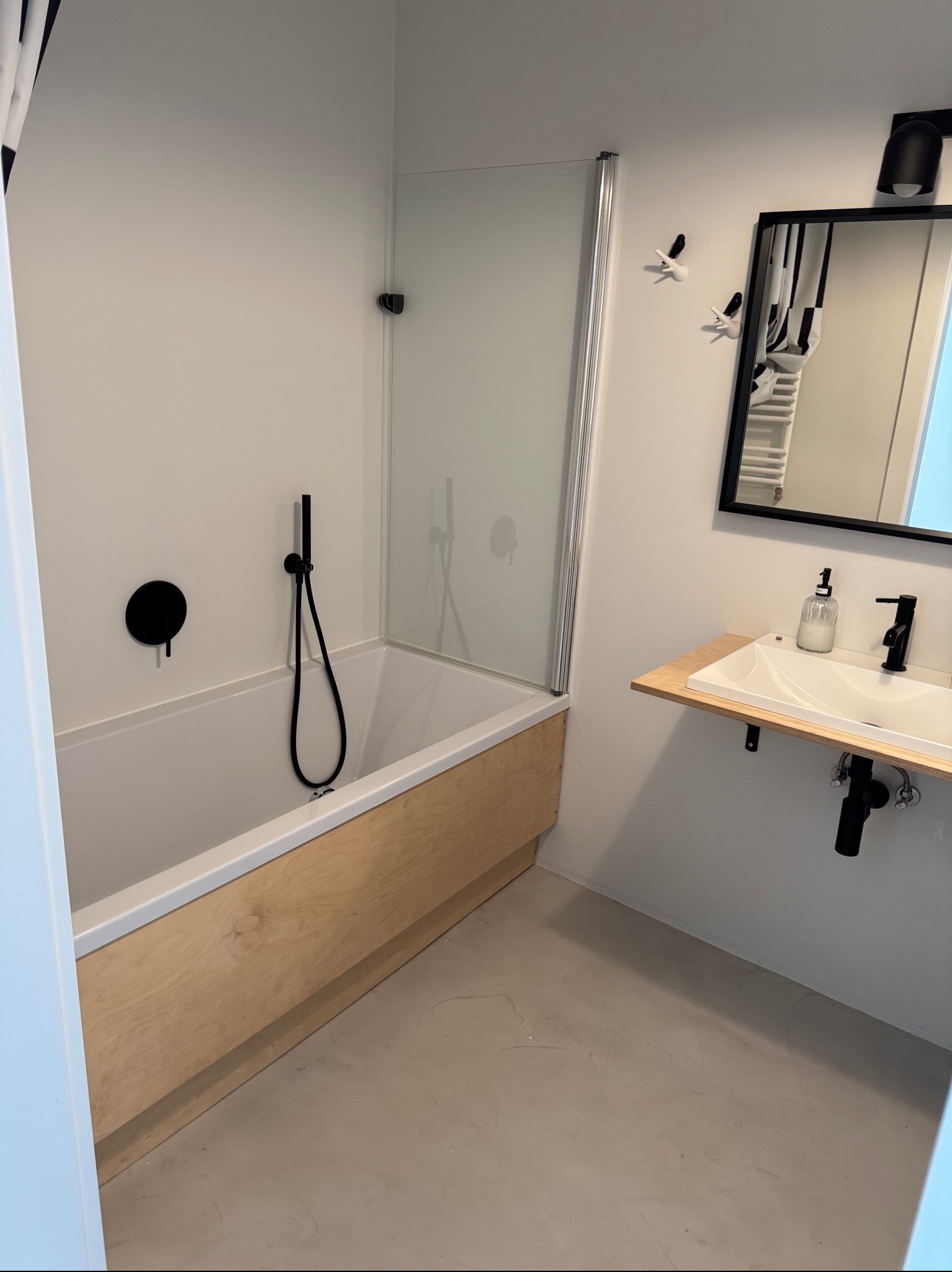 Minimalistyczna łazienka po remoncie: białe ściany, wanna z drewnianą obudową, czarna armatura i lustro w czarnej ramie. Umywalka na drewnianym blacie.