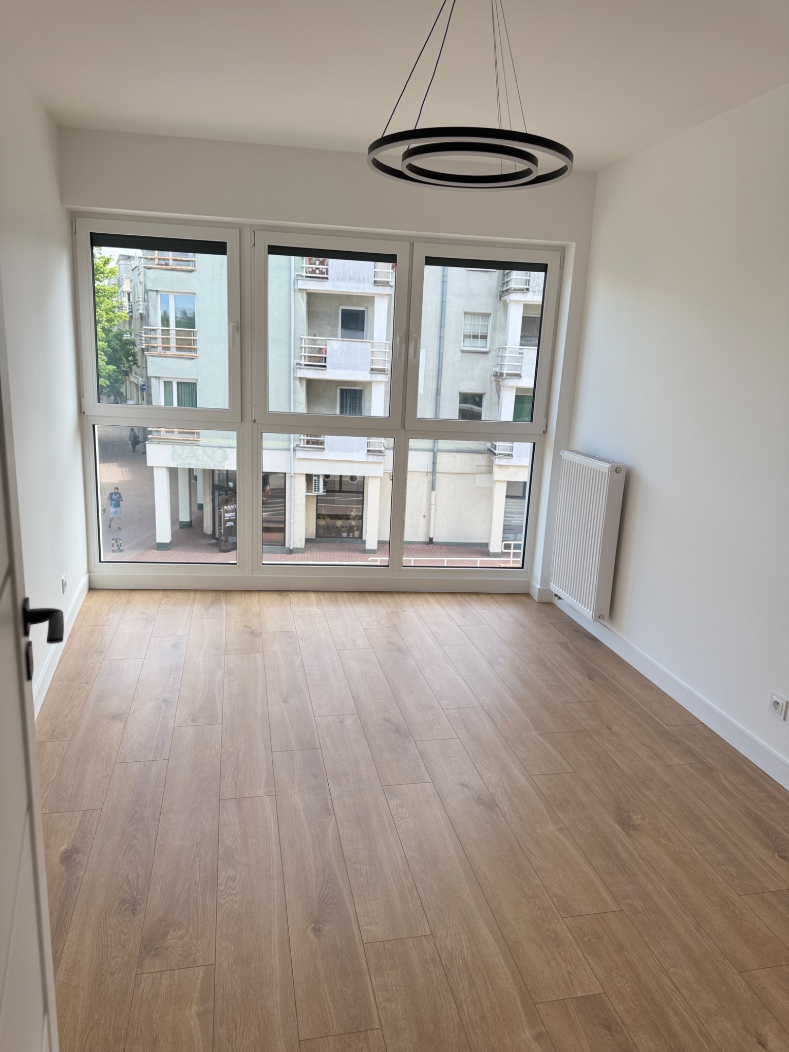 Jasne panele podłogowe w pustym pokoju z białymi ścianami, grzejnikiem i oknem z widokiem na ulicę. Nowoczesna lampa sufitowa.
