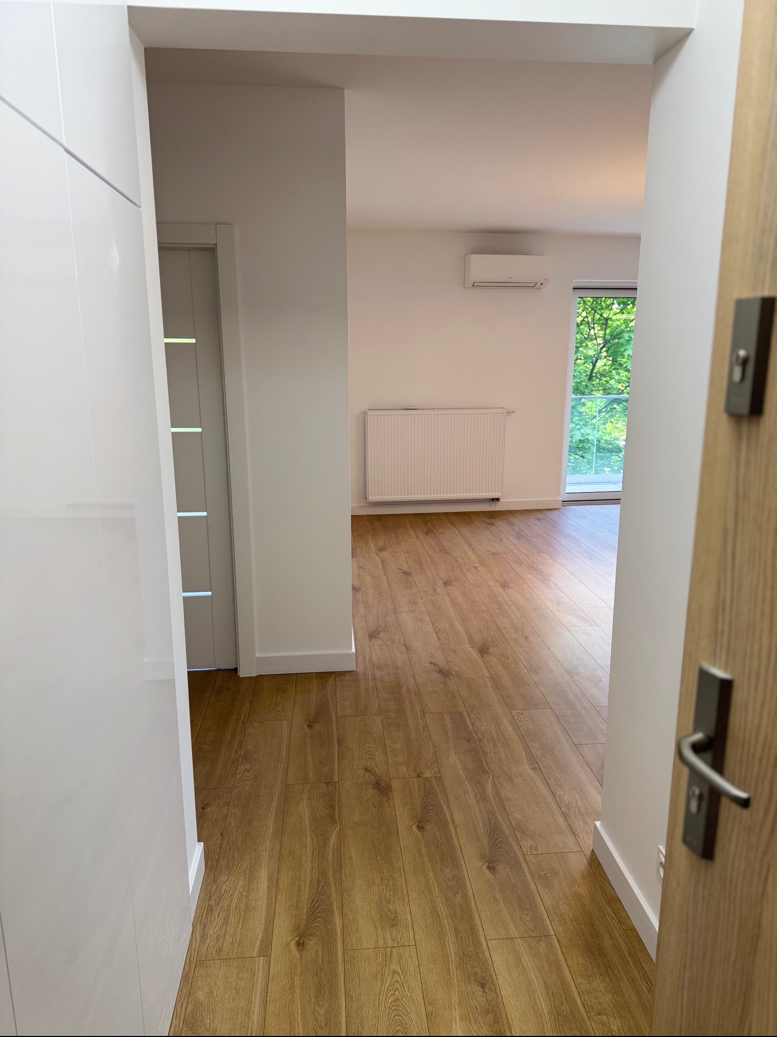 Widok z korytarza na salon z parkietem, grzejnikiem i klimatyzacją. Białe ściany, drzwi drewniane w korytarzu i białe drzwi do pokoju. Jasne wnętrze, widok na zieleń za oknem.