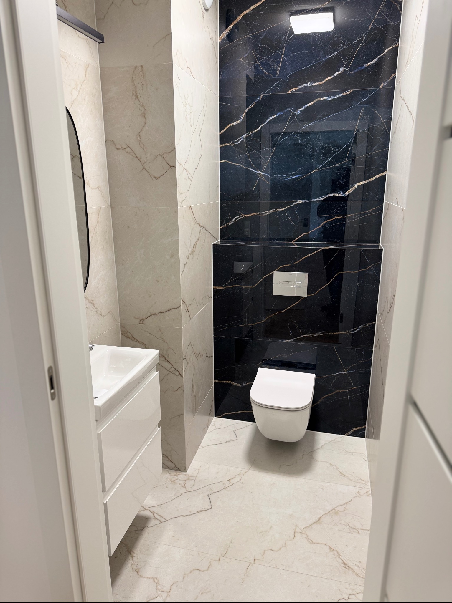 Wykończona łazienka z czarnymi i beżowymi płytkami imitującymi marmur, białą armaturą i szafką pod umywalką. Widok przez otwarte drzwi.