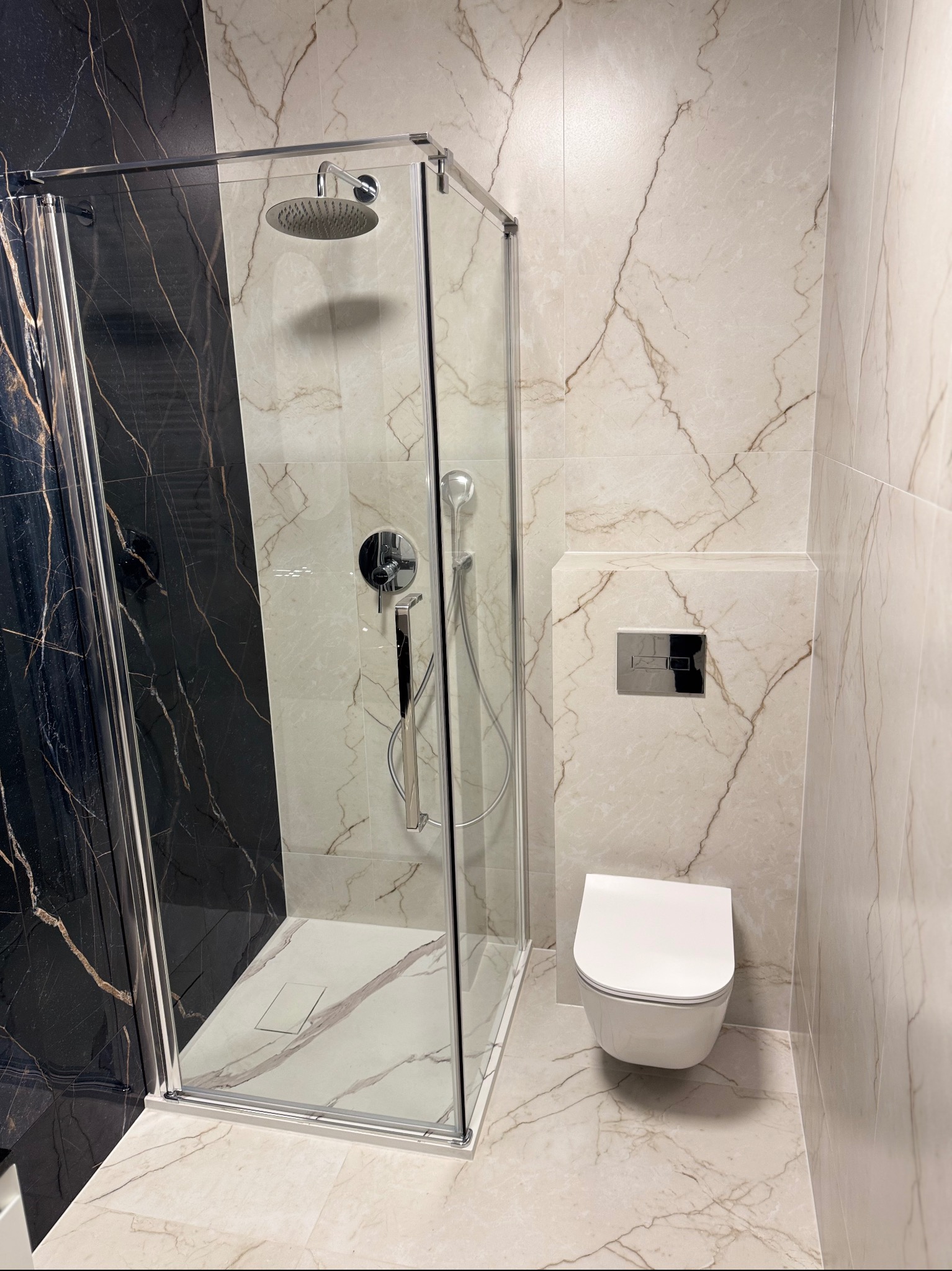 Nowoczesna łazienka z kabiną prysznicową typu walk-in, wykończona płytkami imitującymi marmur w odcieniach bieli i czerni. Widoczna toaleta podwieszana i chromowana armatura.