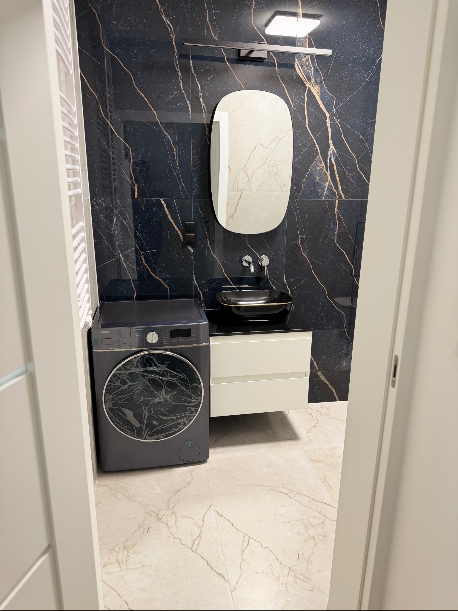 Elegancka łazienka z czarnymi płytkami imitującymi marmur, owalnym lustrem i umywalką nablatową. Pralka w kolorze grafitowym. Całość w nowoczesnym stylu.