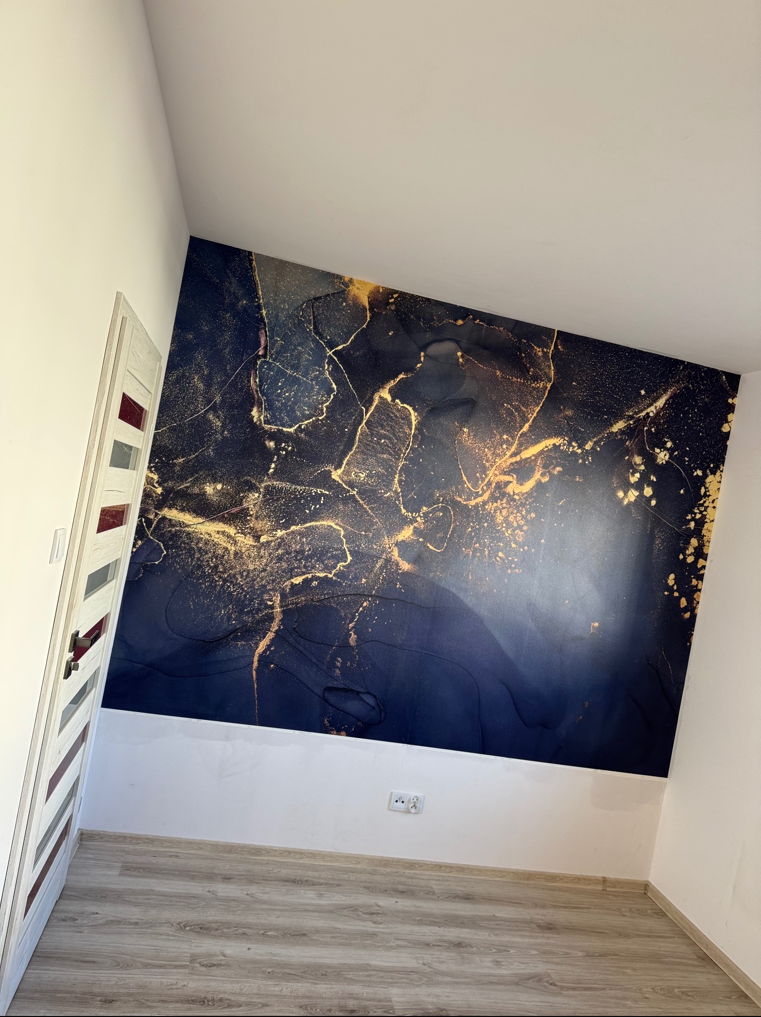 Ściana z abstrakcyjną tapetą w odcieniach granatu i złota w wykończonym pokoju z drewnianą podłogą i białą stolarką drzwiową.