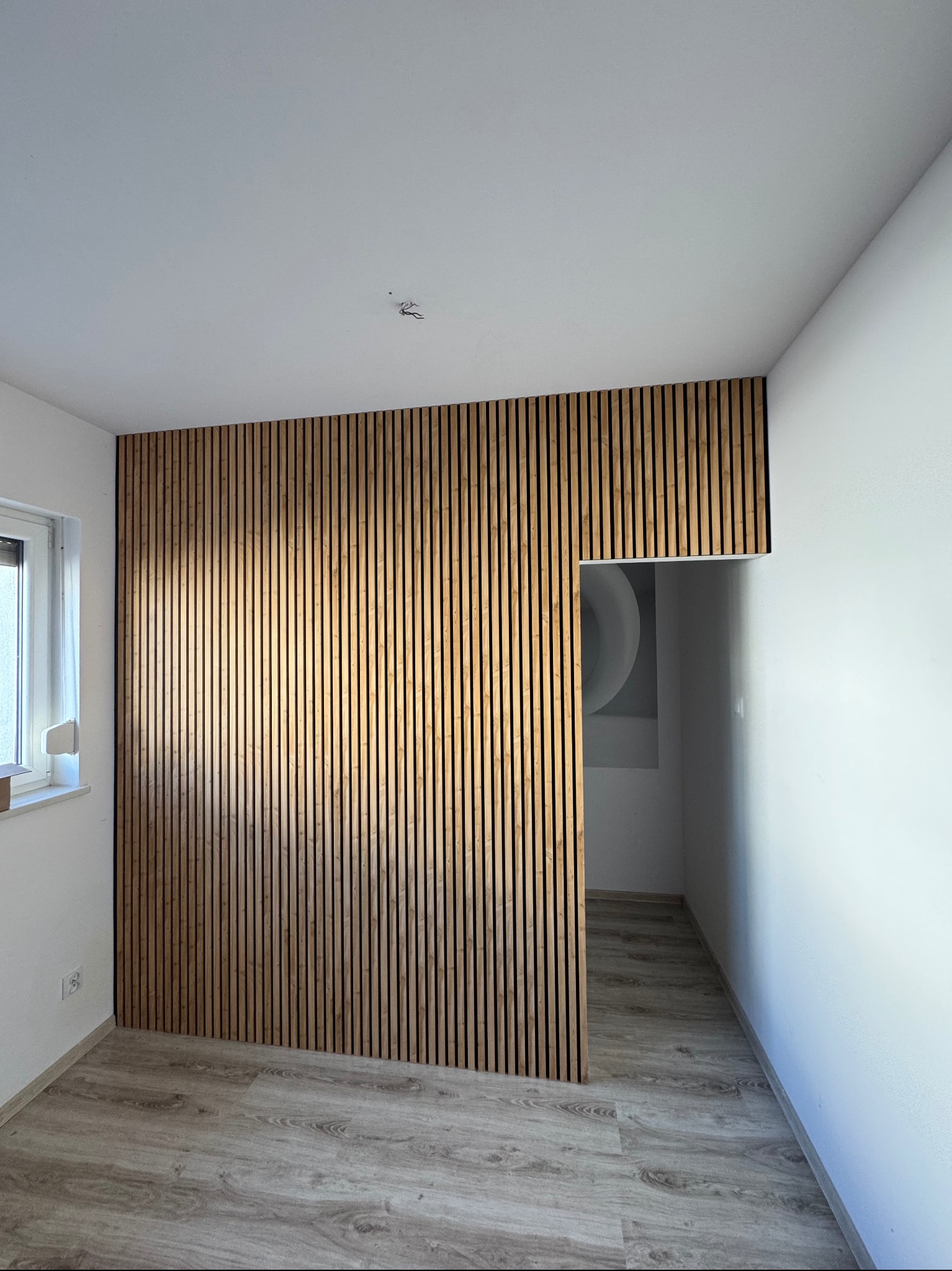 Drewniana ścianka działowa z pionowych lameli, tworząca przejście do innego pomieszczenia. Jasne wnętrze z panelami podłogowymi.
