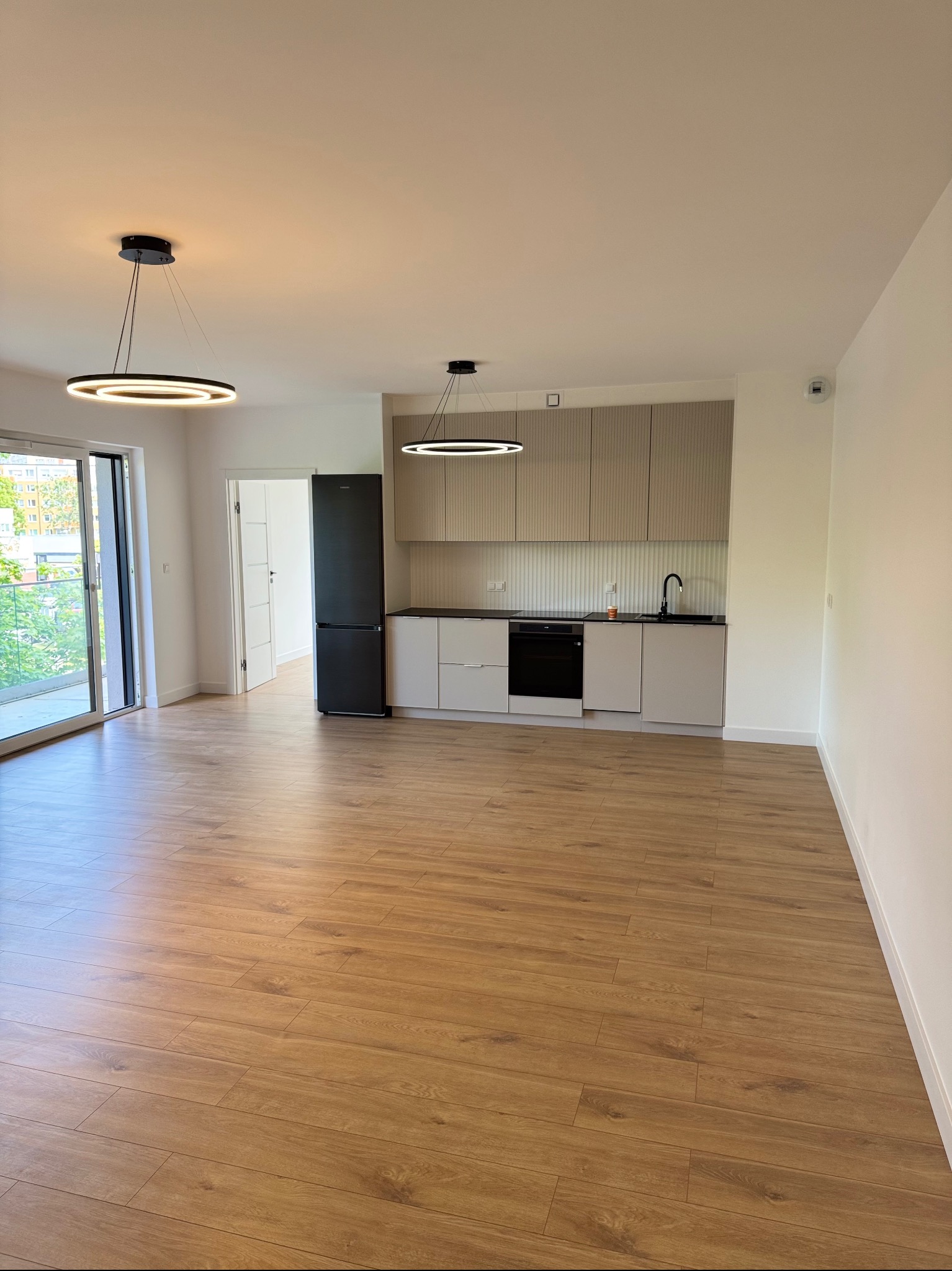 Nowoczesne wnętrze mieszkania z drewnianą podłogą, minimalistyczną kuchnią i czarną lodówką. Widok z balkonu w tle, oświetlenie punktowe.