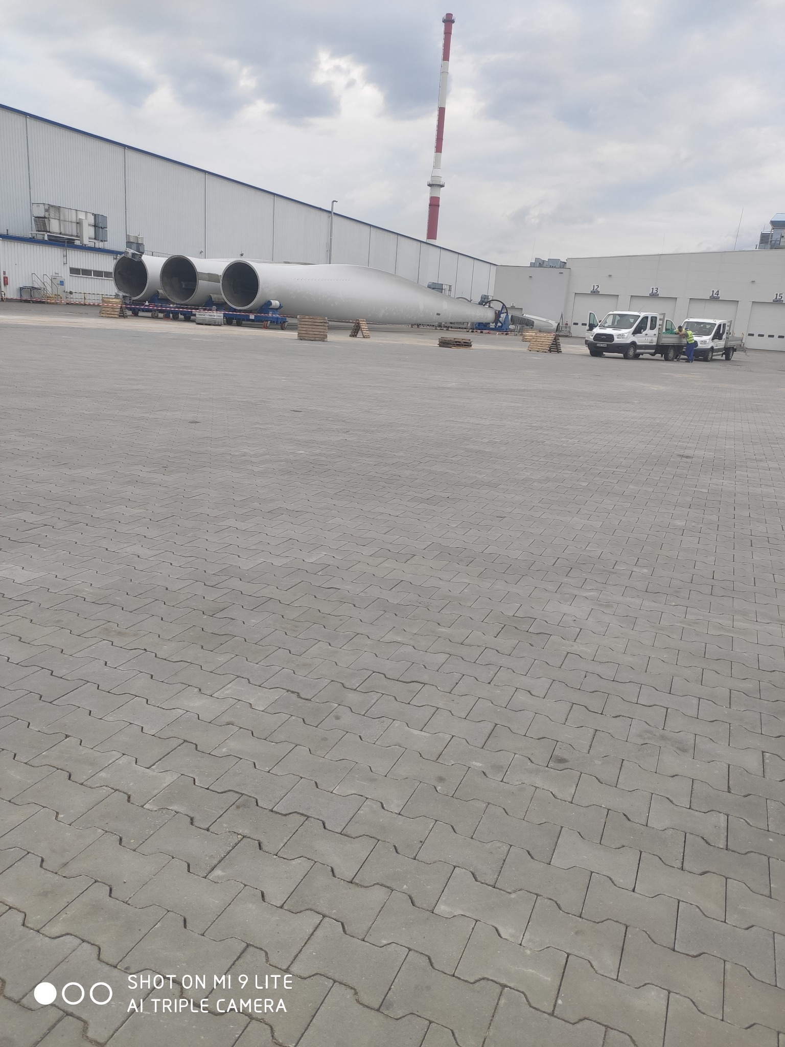 Szeroki plan ułożonej szarej kostki brukowej z widocznymi elementami turbiny wiatrowej w tle, w tym trzy cylindryczne sekcje i jedno skrzydło, oraz dwa samochody dostawcze i komin fabryczny.