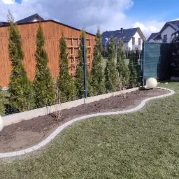 Ogród z równo przystrzyżonym trawnikiem, obrzeżem z kostki granitowej, posadzonymi tujami i lampami kulami, widok na drewnianą altanę i domy w tle.