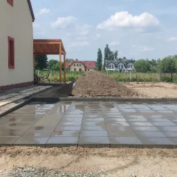 Świeżo ułożona, mokra kostka brukowa na przygotowanym podłożu, widoczny fragment domu z numerem 37, w tle ogród i inne budynki.