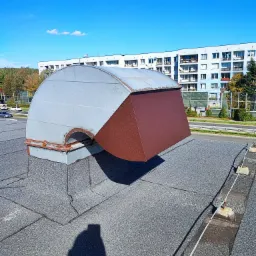 Szara, metalowa obudowa wentylacyjna z brązowym daszkiem na dachu budynku w Jaworznie, widok z góry, w tle budynki mieszkalne i błękitne niebo.