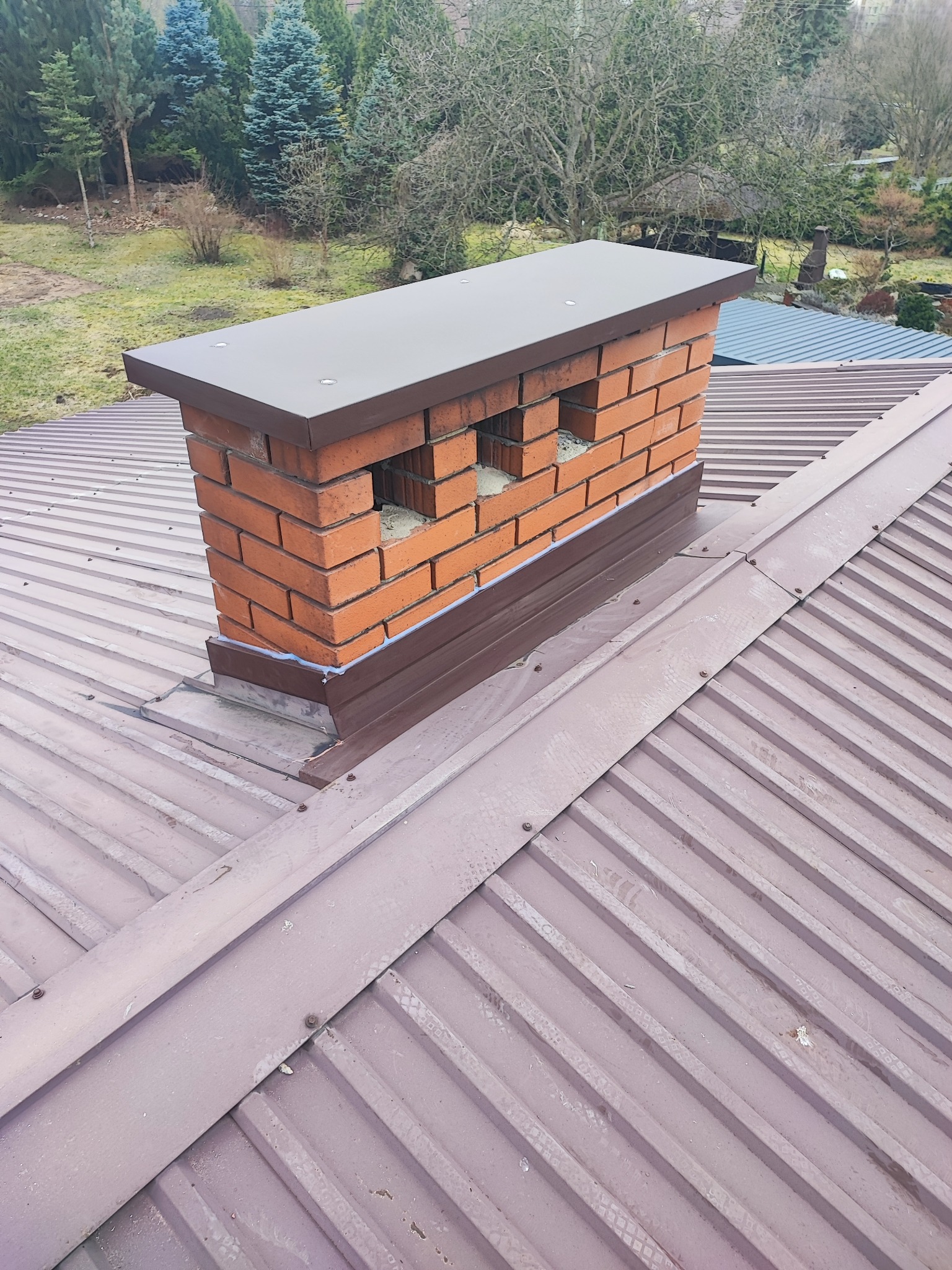 Ceglany komin z brązowym obramowaniem na metalowym dachu w kolorze miedzianym, widok z góry na tle drzew
