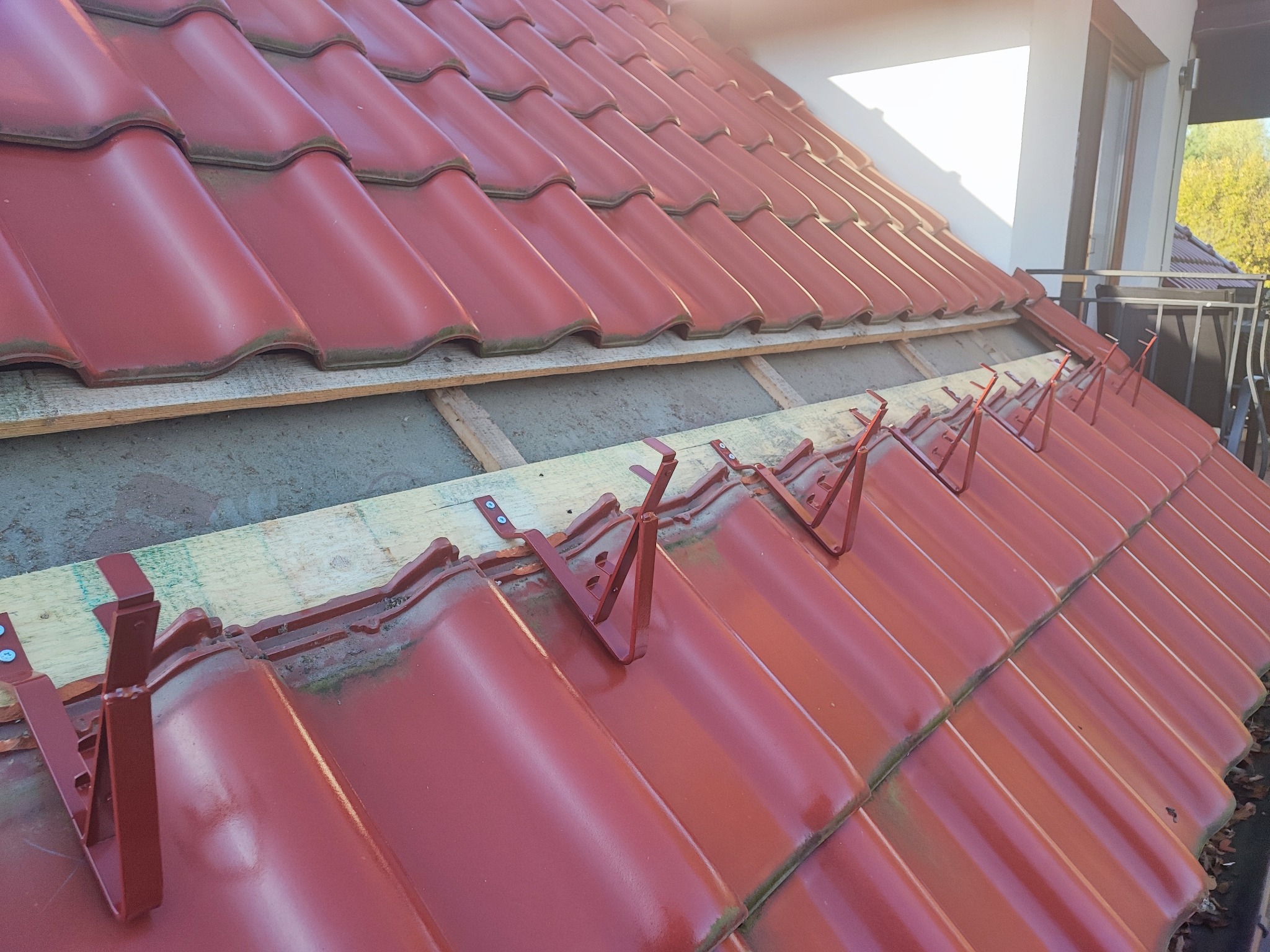 Czerwone dachówki z metalowymi zaczepami na drewnianej łacie, przygotowane do montażu na dachu. Widoczne elementy konstrukcji dachu i fragment budynku w tle.