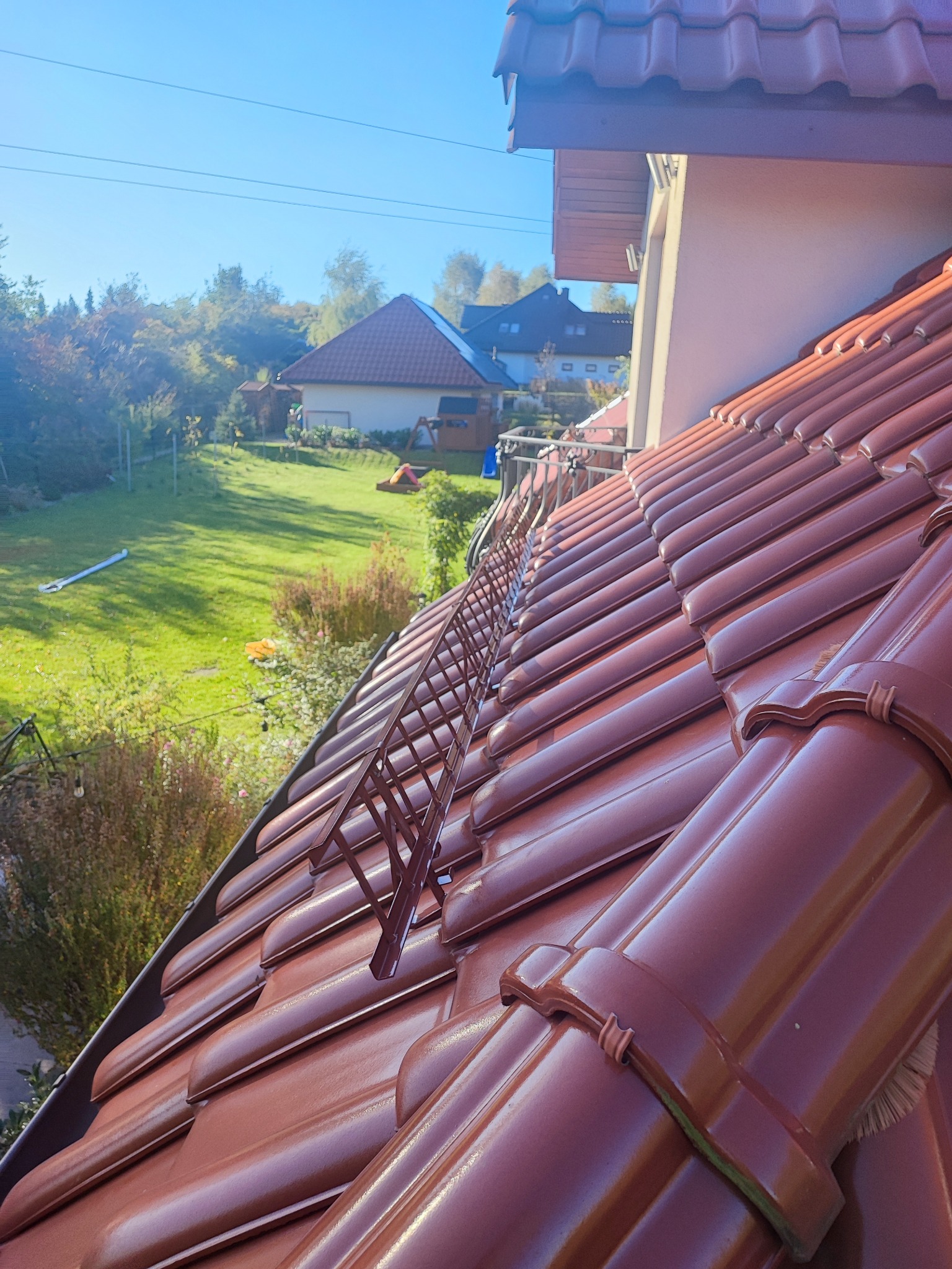 Czerwona dachówka ceramiczna z zamontowanym brązowym stopniem kominiarskim, widok na zielony ogród i domy w tle.