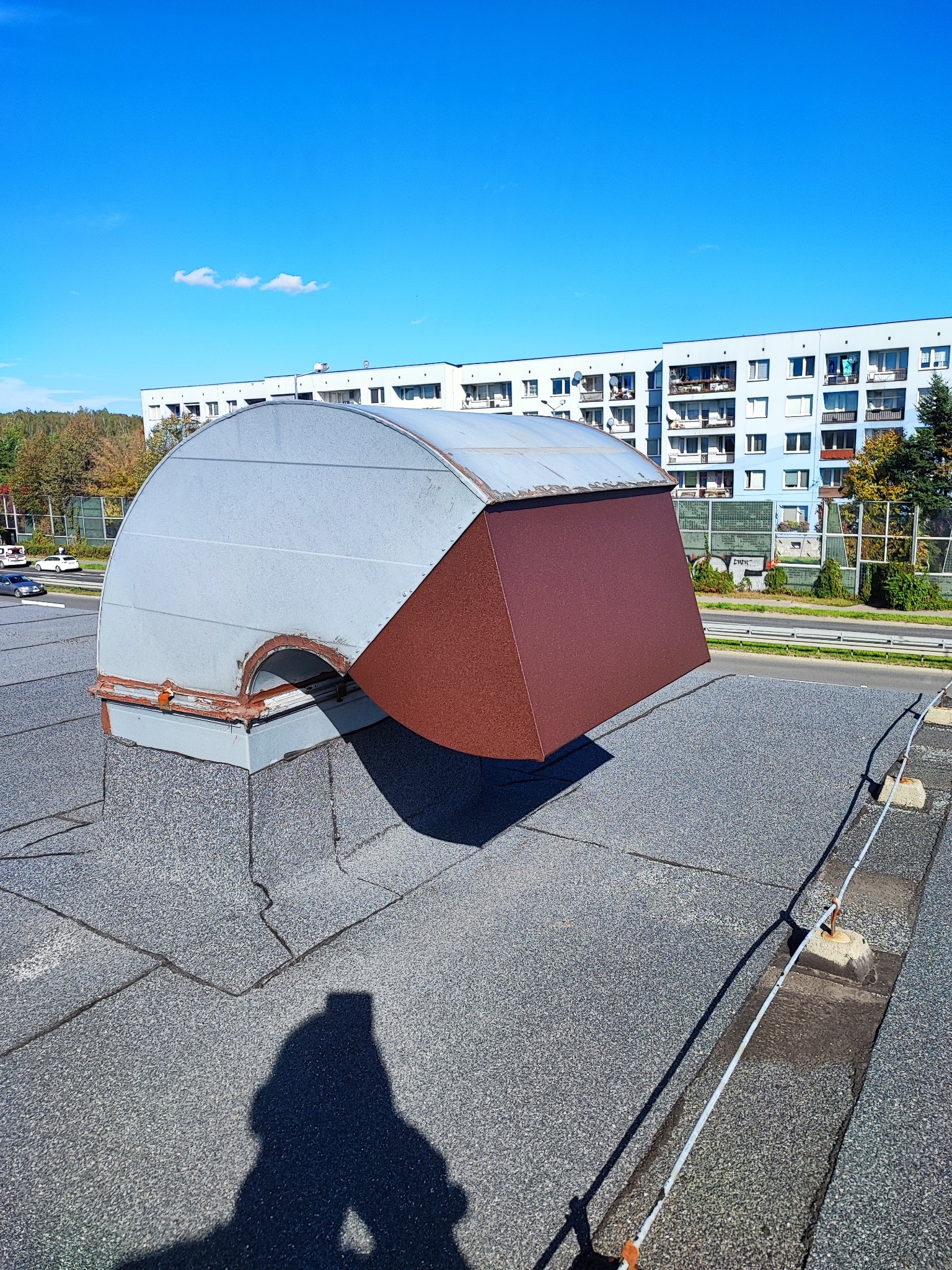 Szara, metalowa obudowa wentylacyjna z brązowym daszkiem na dachu budynku w Jaworznie, widok z góry, w tle budynki mieszkalne i błękitne niebo.