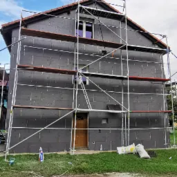 Dwukondygnacyjny dom w trakcie ocieplania elewacji, pokryty czarnym styropianem, otoczony rusztowaniem, z widocznymi workami z materiałami budowlanymi i zabawkami na trawniku.