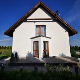 Biała elewacja domu jednorodzinnego z brązowym dachem i oknami, widoczna antena satelitarna i roślinność wokół budynku, ślady po pracach elewacyjnych na podłożu.