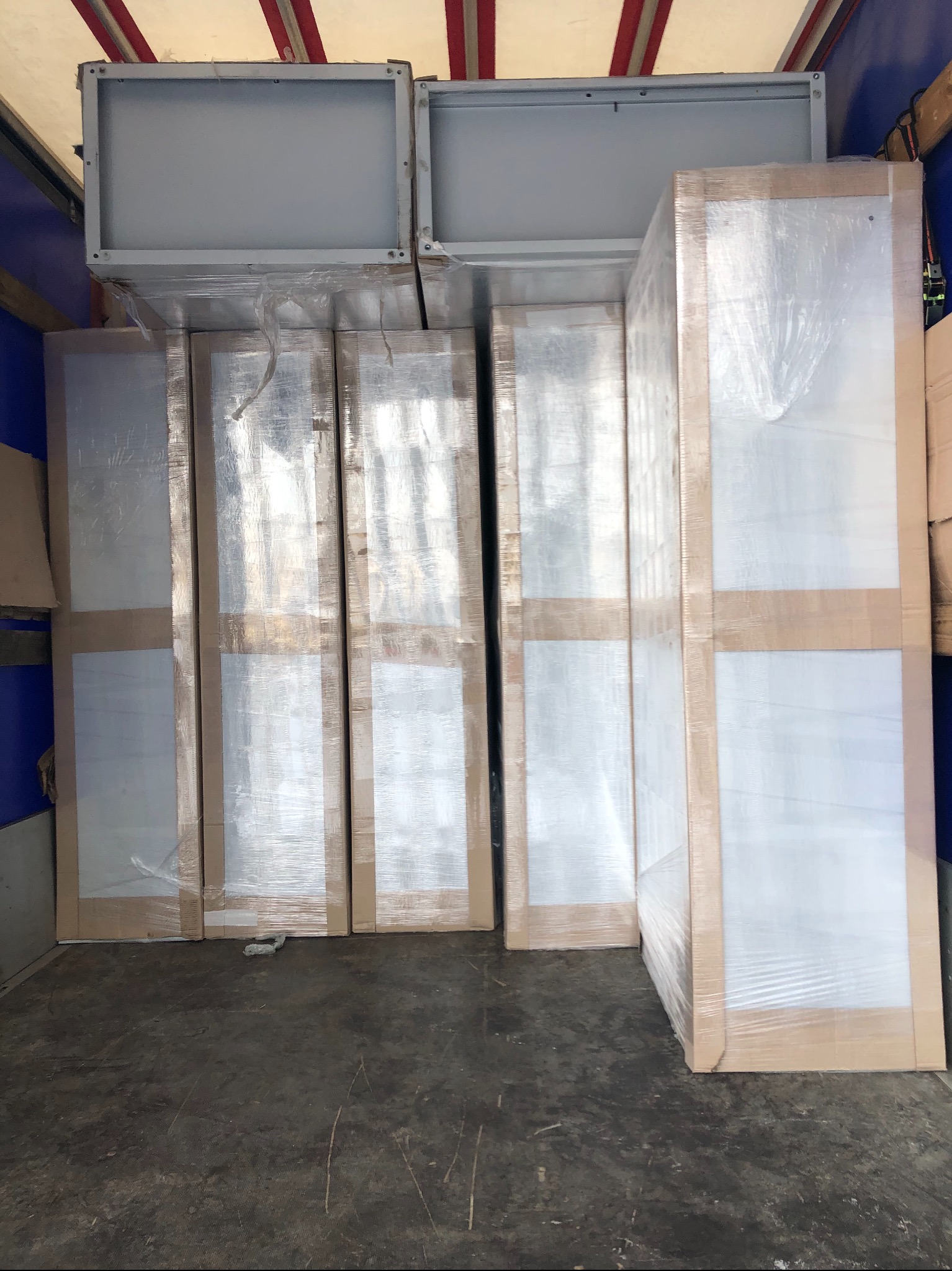 Wnętrze pojazdu transportowego z załadowanymi, zabezpieczonymi folią i kartonem, białymi panelami lub drzwiami, oraz szarymi, metalowymi elementami konstrukcyjnymi