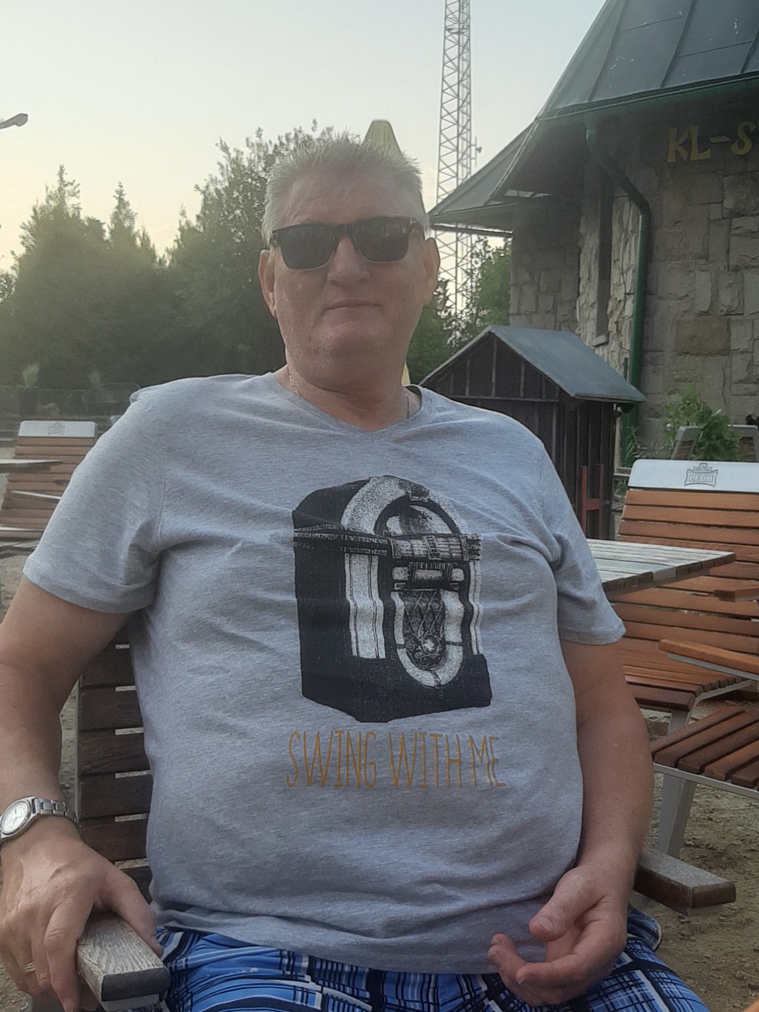 Mężczyzna w okularach przeciwsłonecznych siedzi na drewnianej ławce, ubrany w szarą koszulkę z nadrukiem w stylu jukeboxa i niebieskie szorty w kratę, w tle budynek z kamienia i wieża...