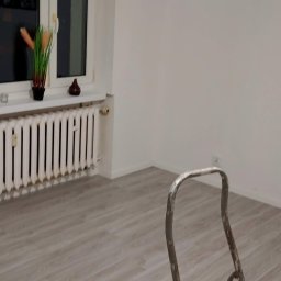 Mirosław Ziemniak - Pokój po odświeżeniu ścian, widoczna drabina malarska, białe ściany, grzejnik i panele podłogowe w odcieniach szarości, lampa sufitowa.