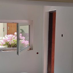 Mirosław Ziemniak - Wnętrze w trakcie malowania: białe ściany, otwarte okno z widokiem na różowe kwiaty na zewnątrz, widoczne ślady po szpachlowaniu i przygotowaniu do malowania.