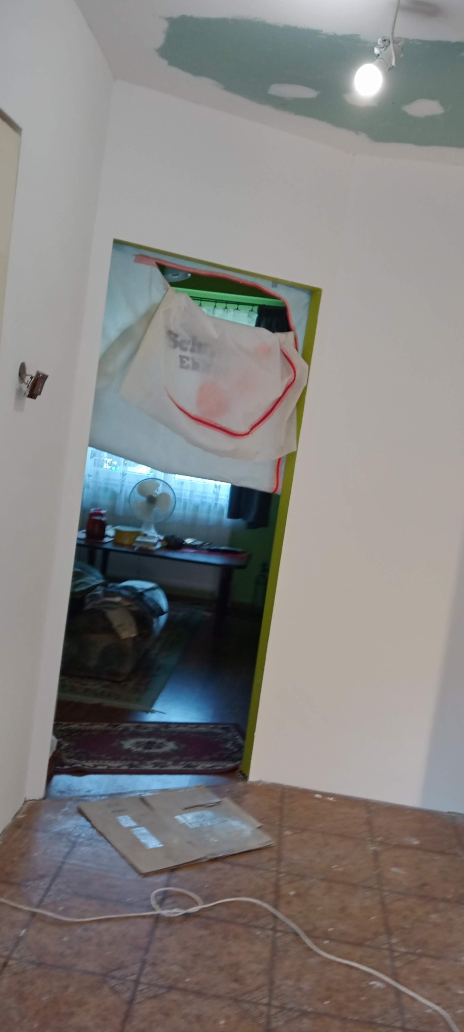 Pokój w trakcie remontu: świeżo pomalowane ściany na biało, sufit z widocznymi śladami szpachlowania, podłoga zabezpieczona kartonem, widok na pokój obok z wentylatorem i dywanem.