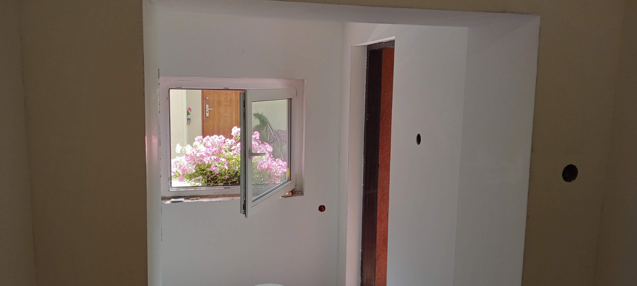 Wnętrze w trakcie malowania: białe ściany, otwarte okno z widokiem na różowe kwiaty na zewnątrz, widoczne ślady po szpachlowaniu i przygotowaniu do malowania.