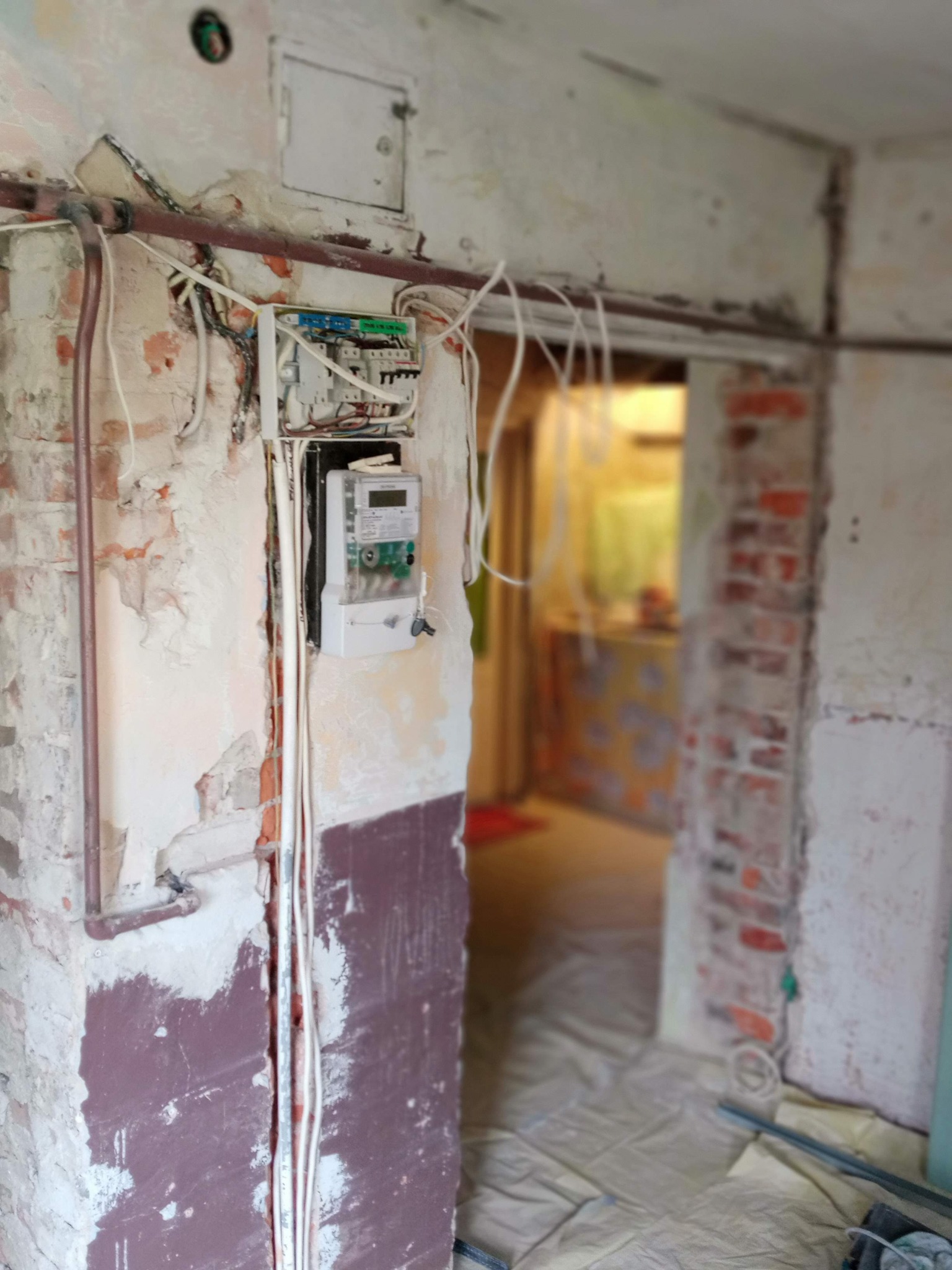 Ściana w trakcie remontu z odsłoniętym licznikiem elektrycznym i przewodami, fragment ściany pomalowany na fioletowo, widoczne ślady po starych instalacjach i fragment muru z cegły w tle.