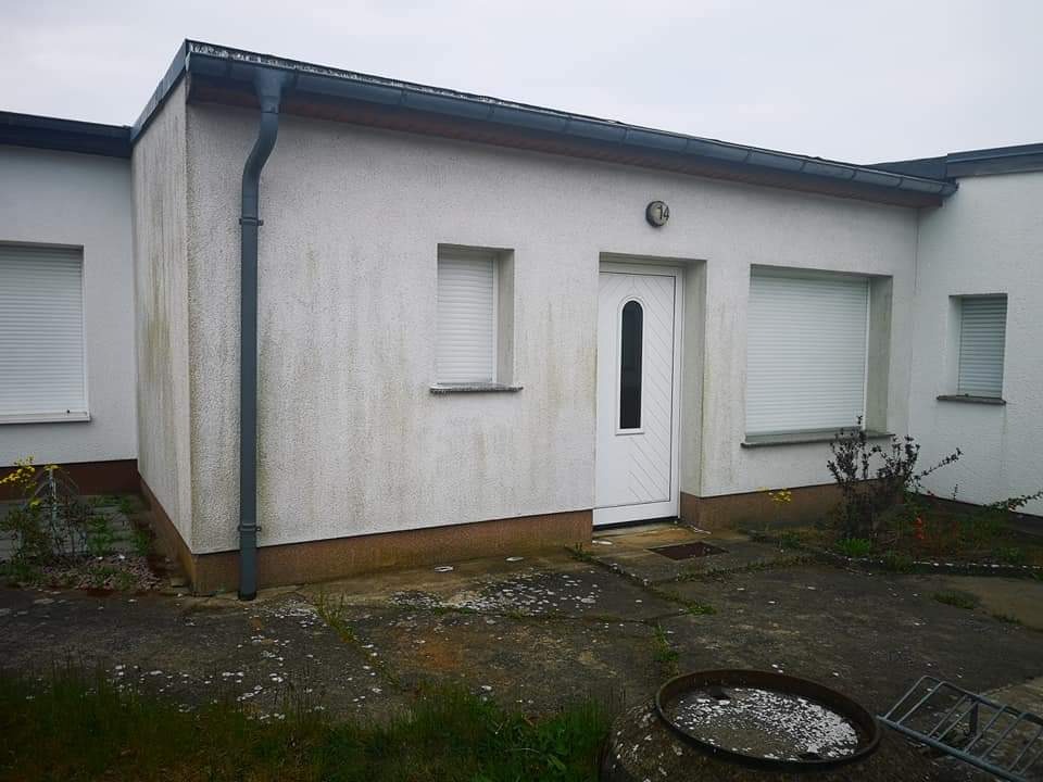 Biała elewacja budynku z widocznymi zabrudzeniami i zaciekami, szara rynna pionowa, białe rolety okienne, białe drzwi wejściowe z numerem 14 nad nimi, zaniedbany chodnik z resztkami roślinności.