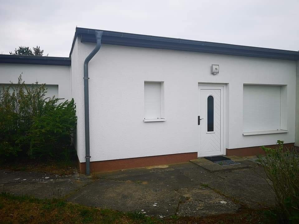 Biały, parterowy budynek z szarym orynnowaniem i białymi roletami zewnętrznymi, widok z zewnątrz w pochmurny dzień.