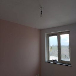 MGS-BUD Remonty i Wykończenia - Pomieszczenie po malowaniu: ściana w odcieniach różu, biały sufit z żarówką, okno z widokiem na zewnątrz i puszka farby na parapecie. Podłoga drewniana.