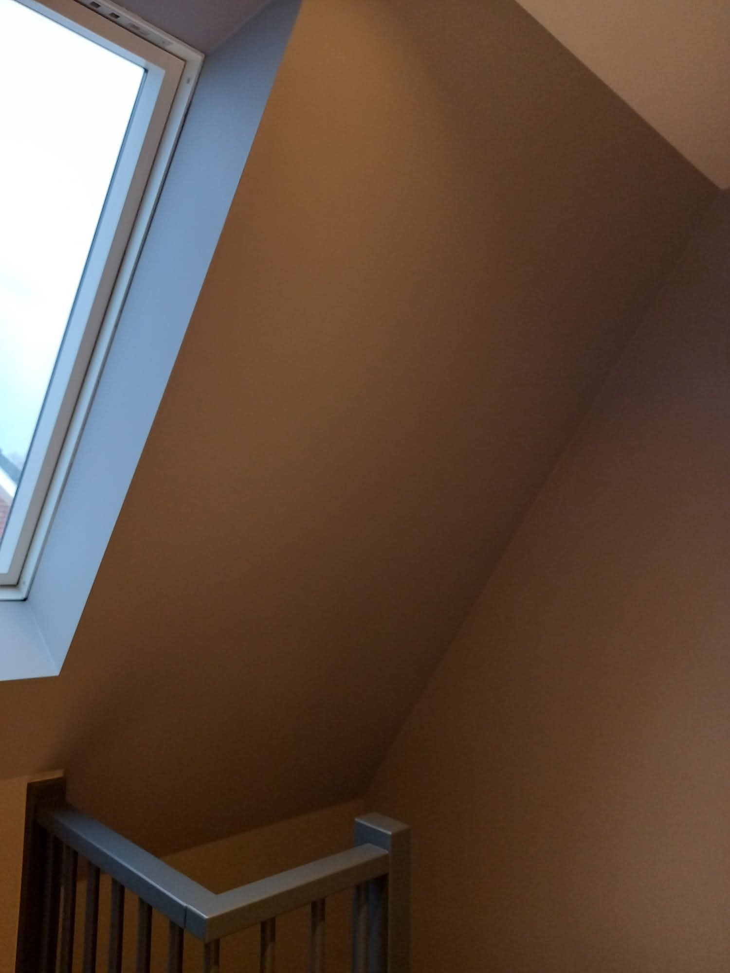 Pomarańczowa ściana na poddaszu z oknem dachowym i fragmentem szarej balustrady schodów. Ściana świeżo pomalowana, bez smug i zacieków.