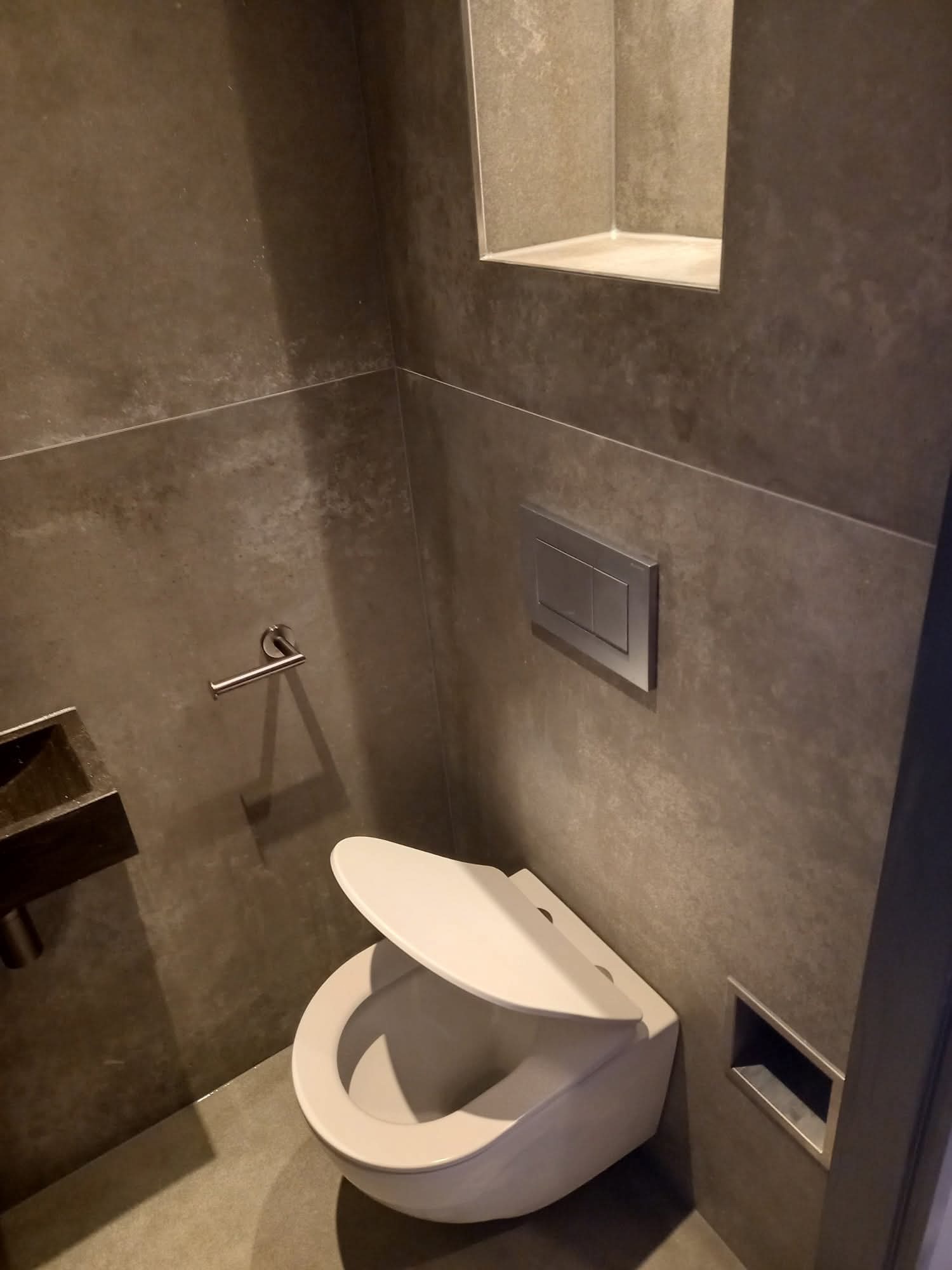 Nowoczesna łazienka z szarymi płytkami, podwieszaną toaletą z otwartą klapą i wnęką w ścianie. Minimalistyczny design, metalowy przycisk spłukiwania.