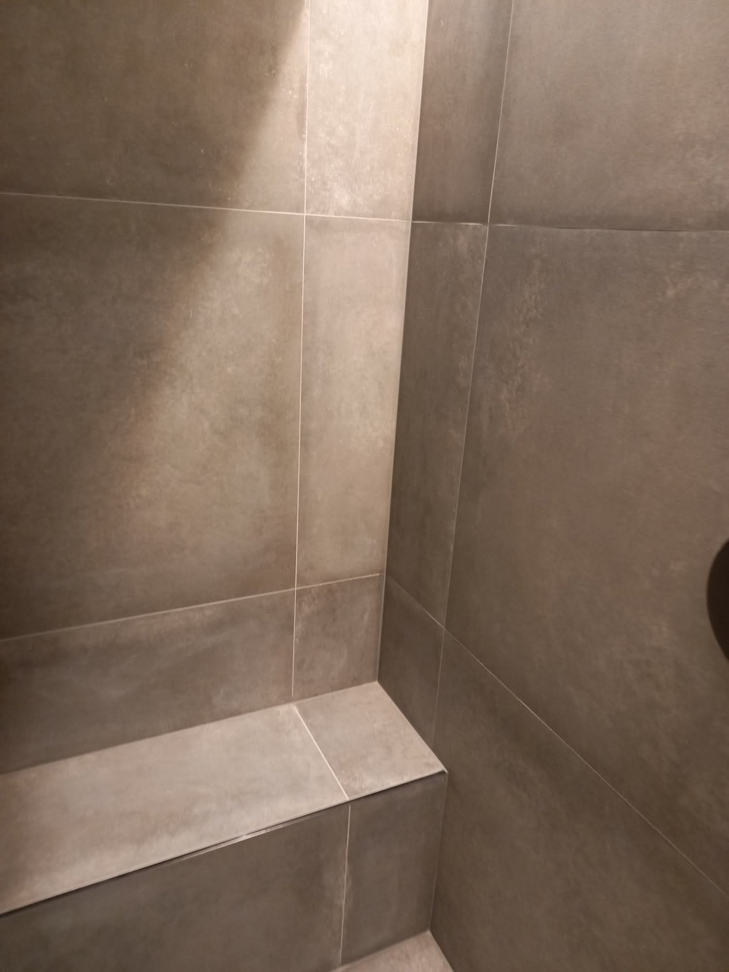 Minimalistyczna łazienka z szarymi, matowymi płytkami wielkoformatowymi i wbudowanym siedziskiem. Czyste linie, stonowana kolorystyka, nowoczesny design.