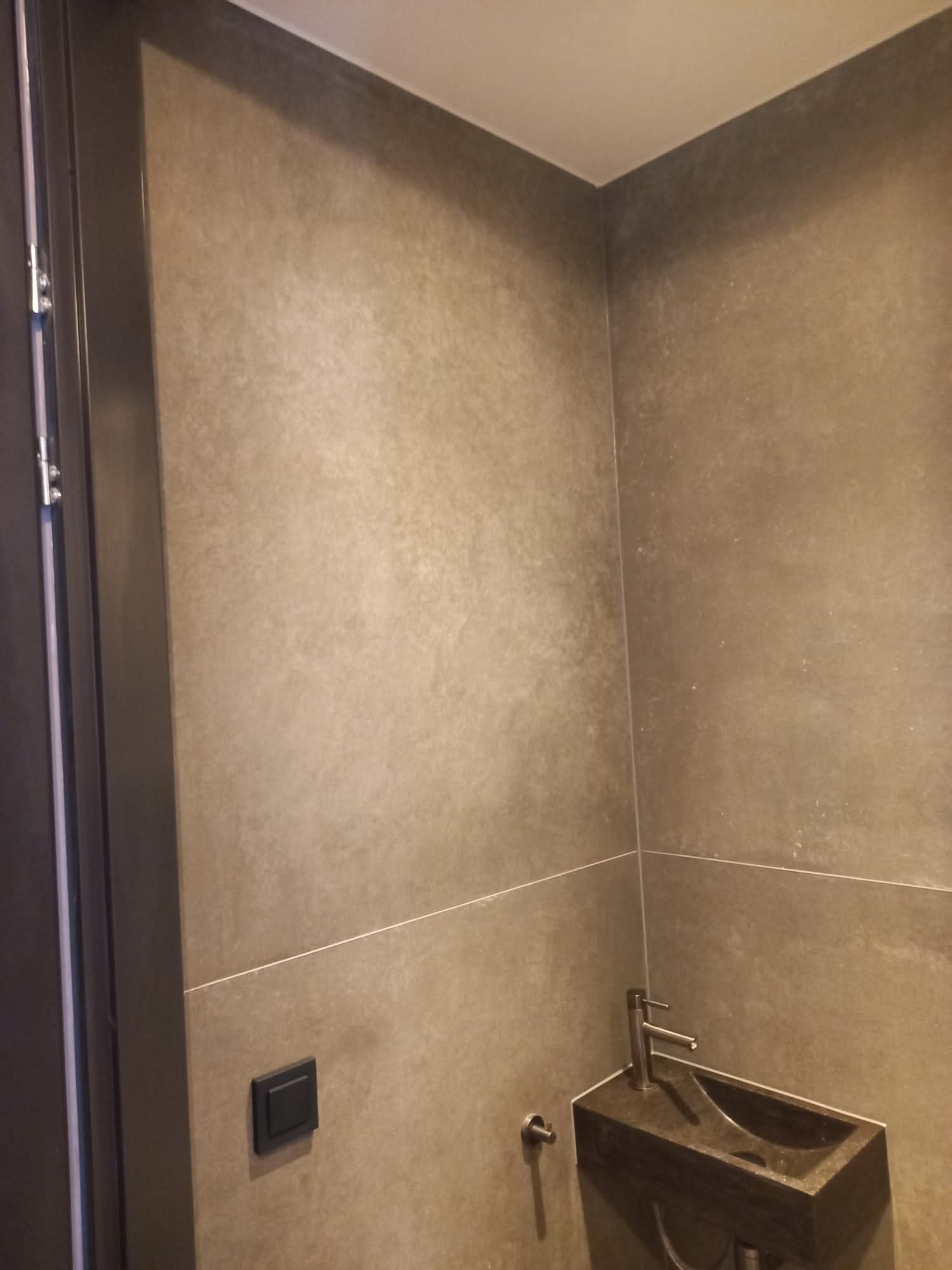 Wykończona łazienka z umywalką z kamienia i szarą, teksturowaną okładziną ścienną. Widoczny fragment drzwi i włącznik światła.