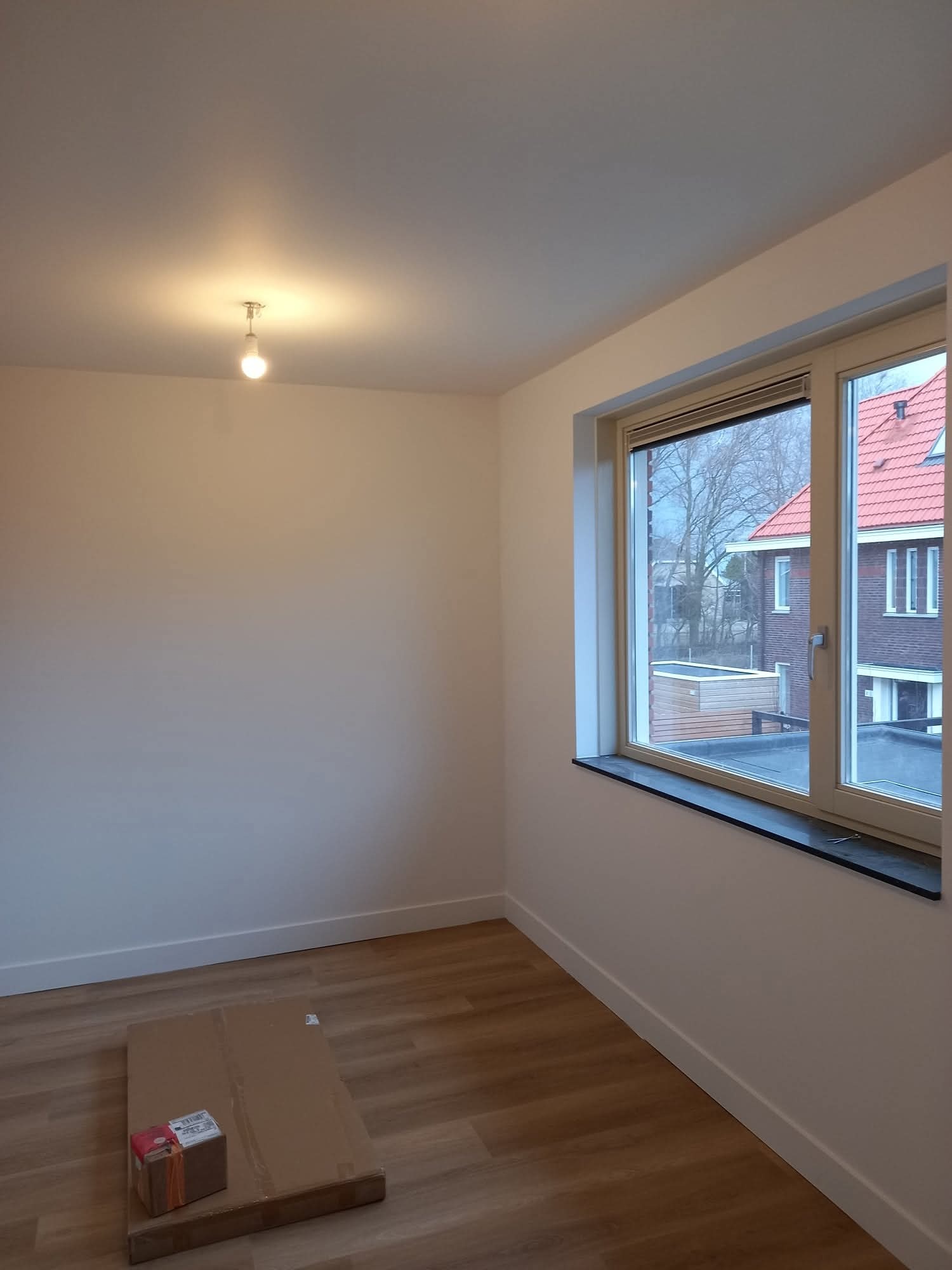 Pomalowane na biało wnętrze z drewnianą podłogą i oknem. Na podłodze leżą kartony. Widok na budynek z czerwonym dachem za oknem.