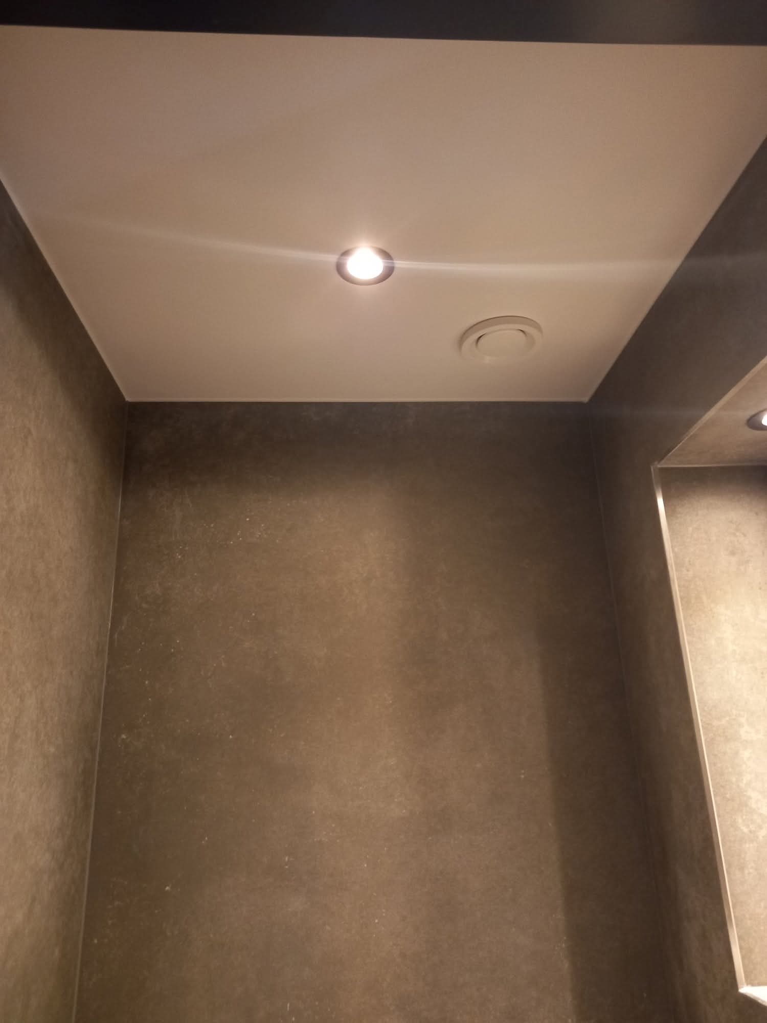 Wykończona łazienka z szarobrązowymi ścianami, podwieszanym sufitem z oświetleniem punktowym i kratką wentylacyjną. Minimalistyczny design.