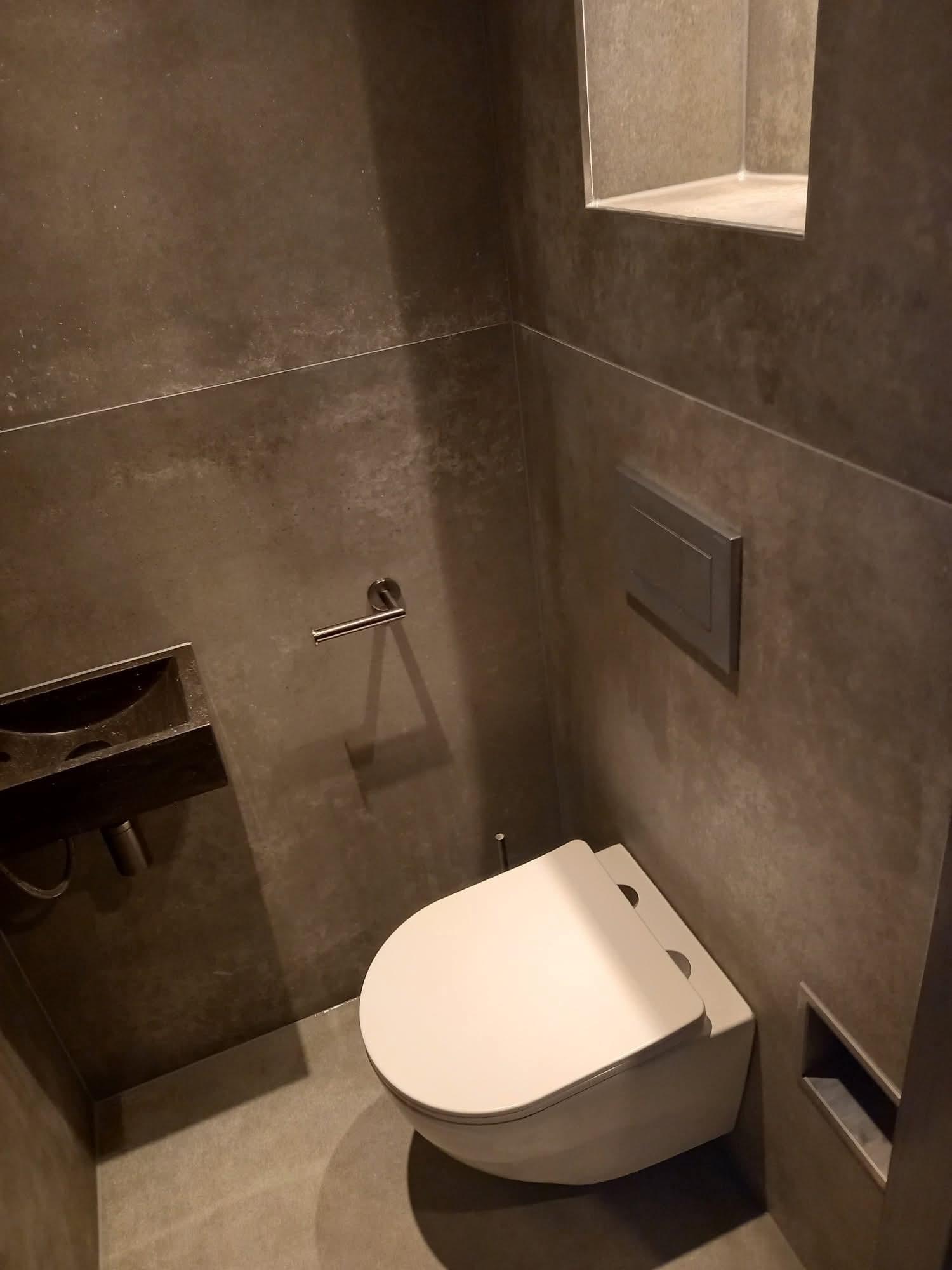 Nowoczesna łazienka w odcieniach szarości z podwieszaną toaletą, minimalistyczną umywalką i wnęką w ścianie. Wykończenie ścian imituje beton.