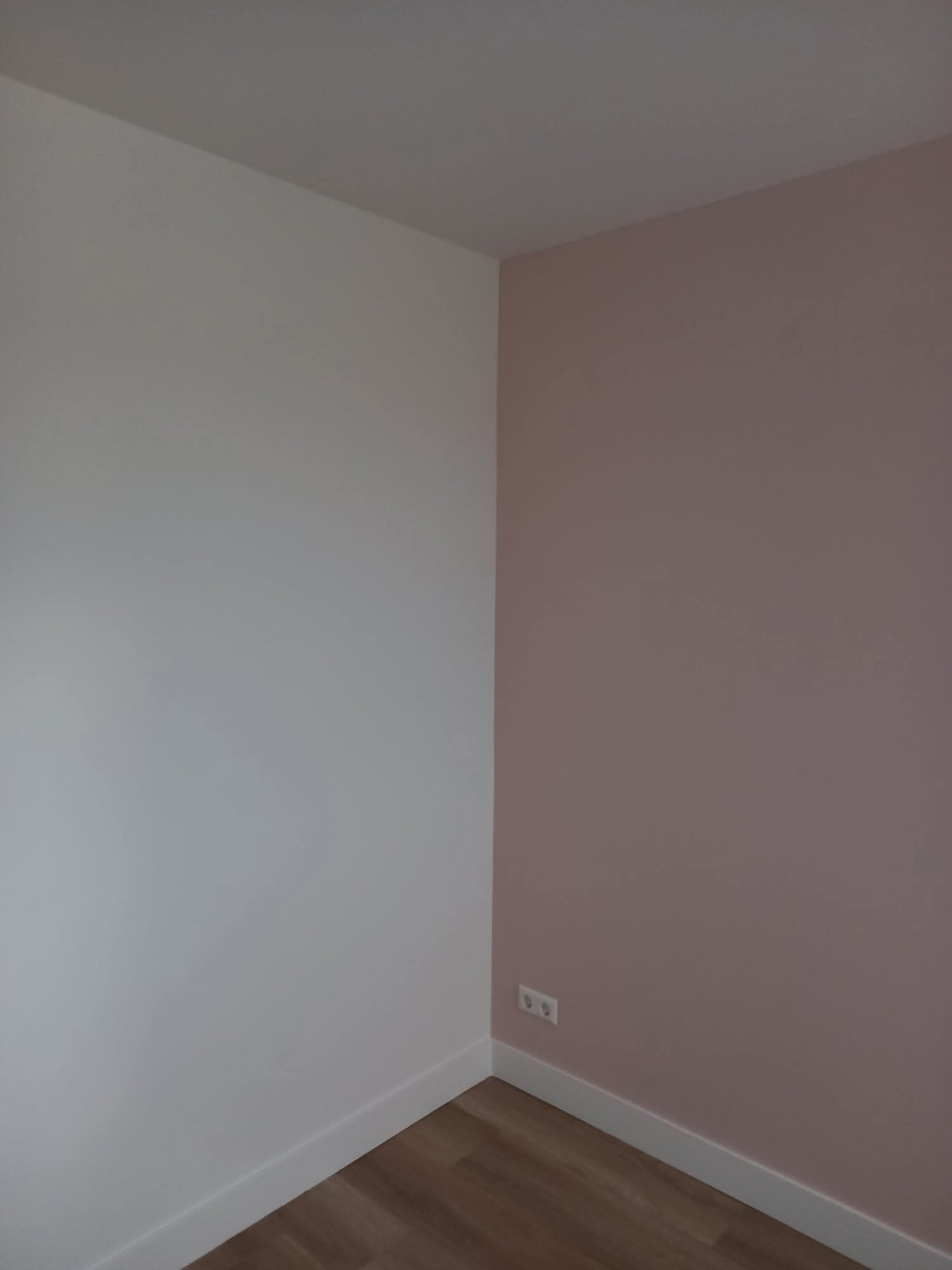 Pomieszczenie po malowaniu: ściana biała i różowa, listwy przypodłogowe, panele podłogowe. Widoczne gniazdka elektryczne. Minimalistyczny design.