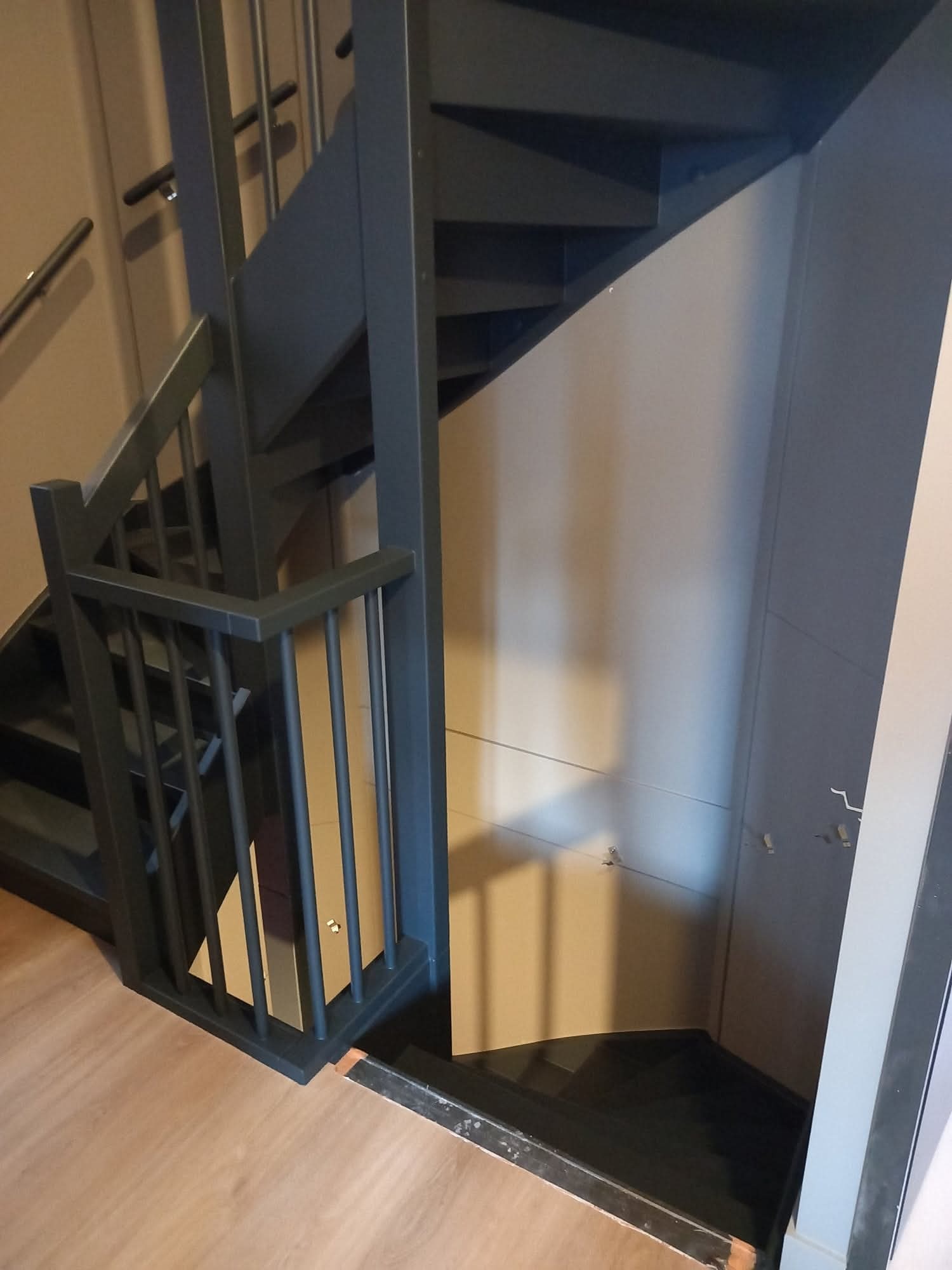 Nowoczesne, ciemne schody drewniane z metalowymi balustradami, prowadzące w dół. Widoczne detale wykończenia i połączenie z podłogą.