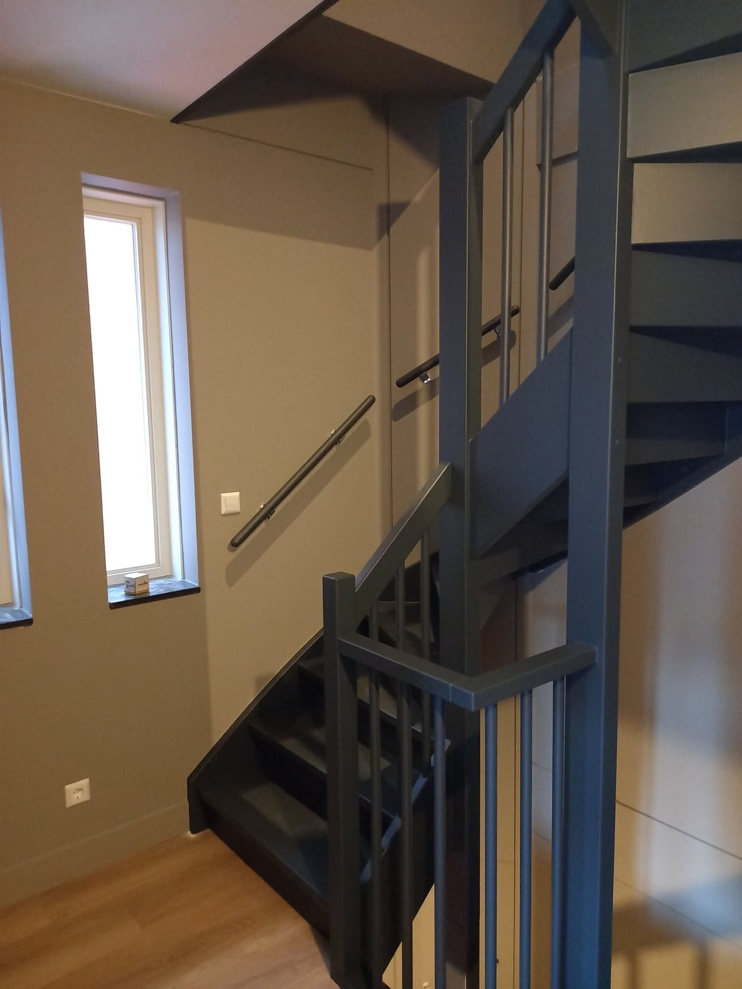 Nowoczesne, ciemne schody z metalową balustradą w jasnym wnętrzu. Widoczne okno i drewniana podłoga. Minimalistyczny design.