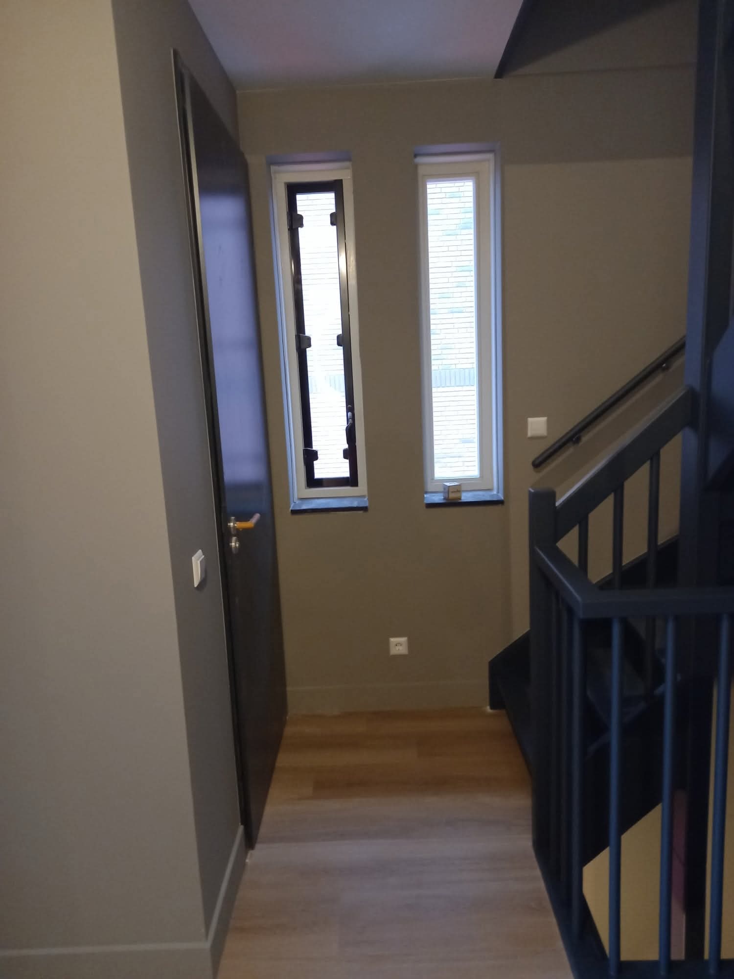 Korytarz po remoncie: szare ściany, drewniana podłoga, czarne drzwi i schody. Dwa wąskie okna z żaluzjami wpuszczają światło.